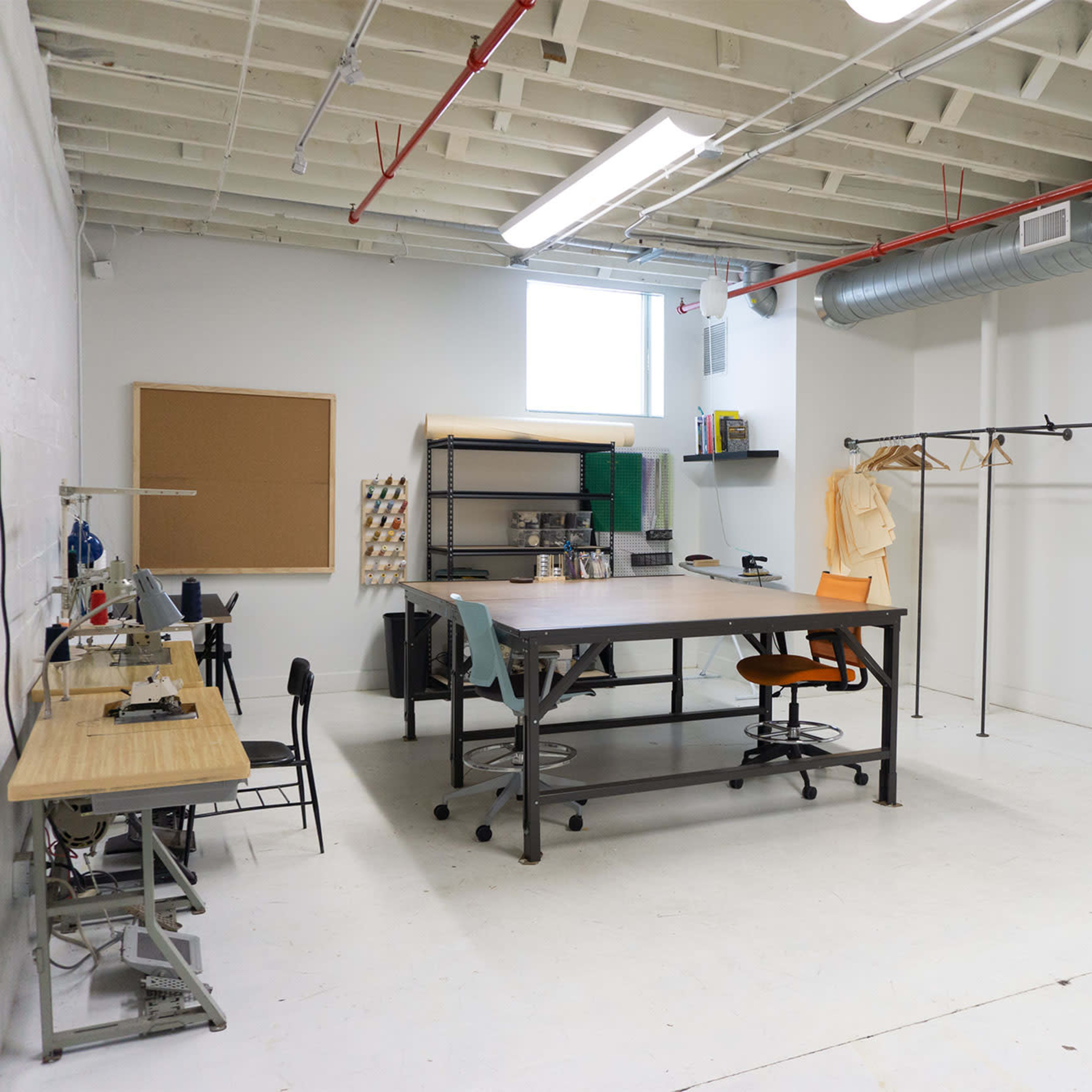 A spacious, industrial-style office workspace with a large central table, chairs, a clothes rack, and a mirror against a white wall.