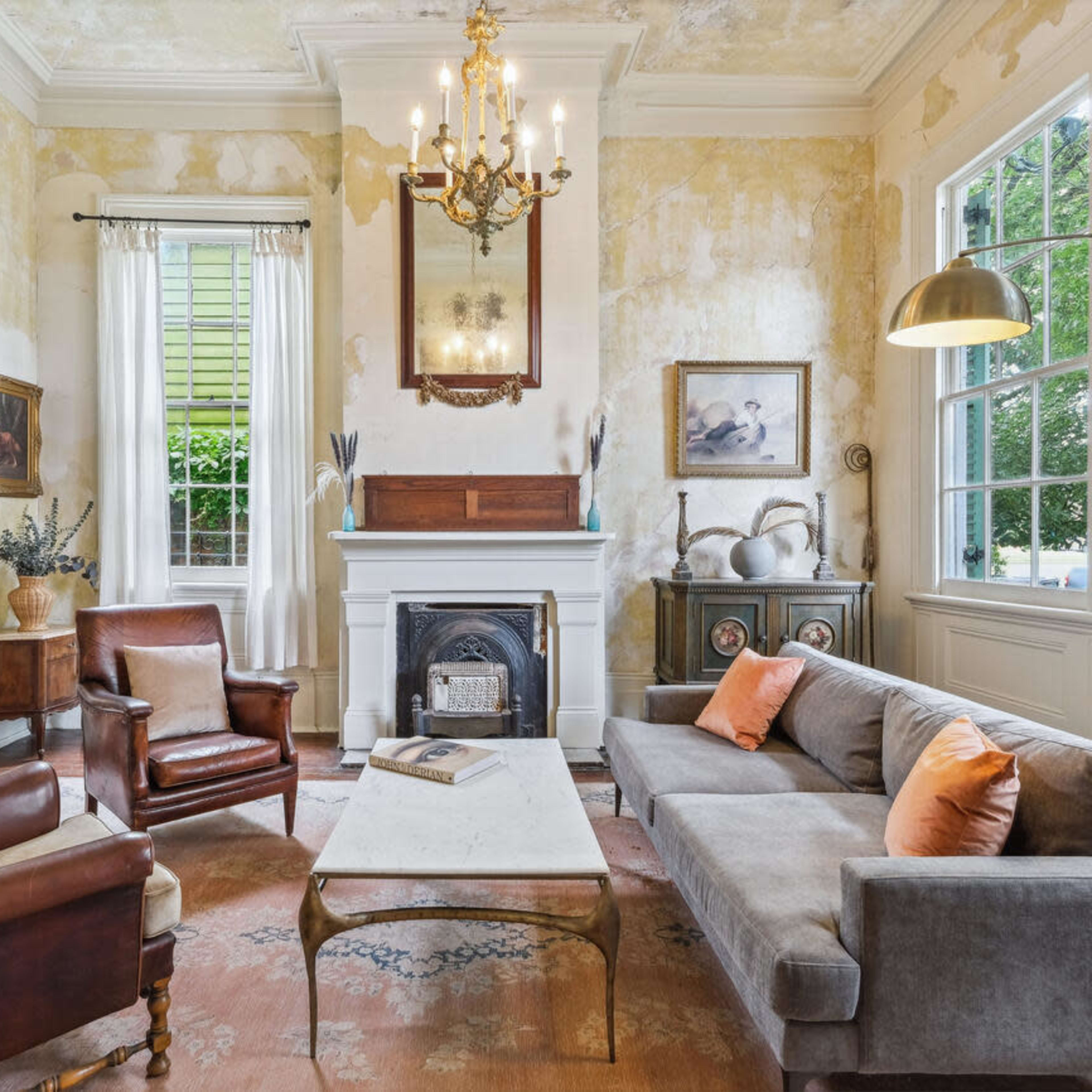 A living room with a chandelier, a fireplace, two armchairs, and a sofa, surrounded by faded wallpaper and large windows.