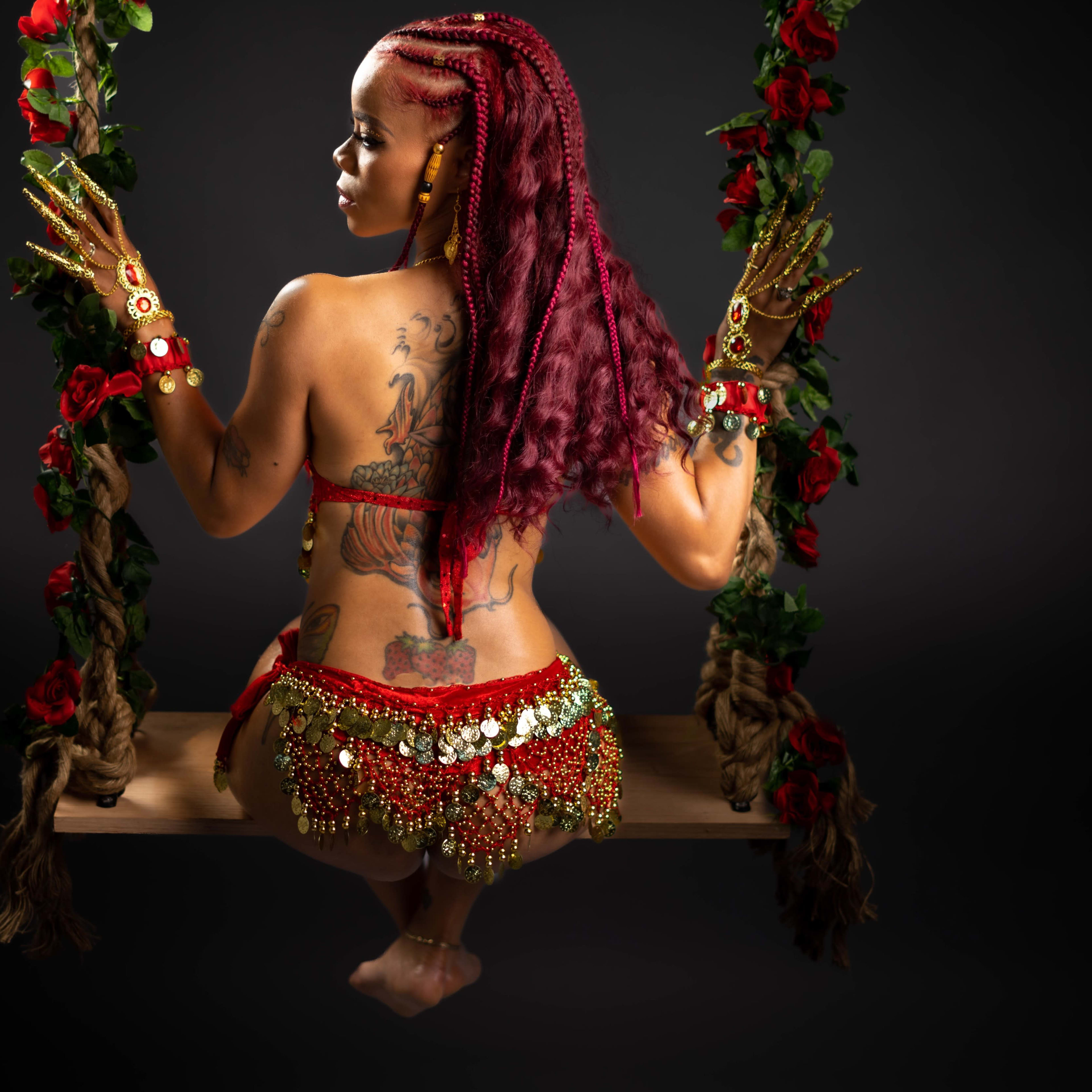 A woman with braided hair and intricate tattoos sits on a swing adorned with flowers, wearing a decorative outfit with sequins.