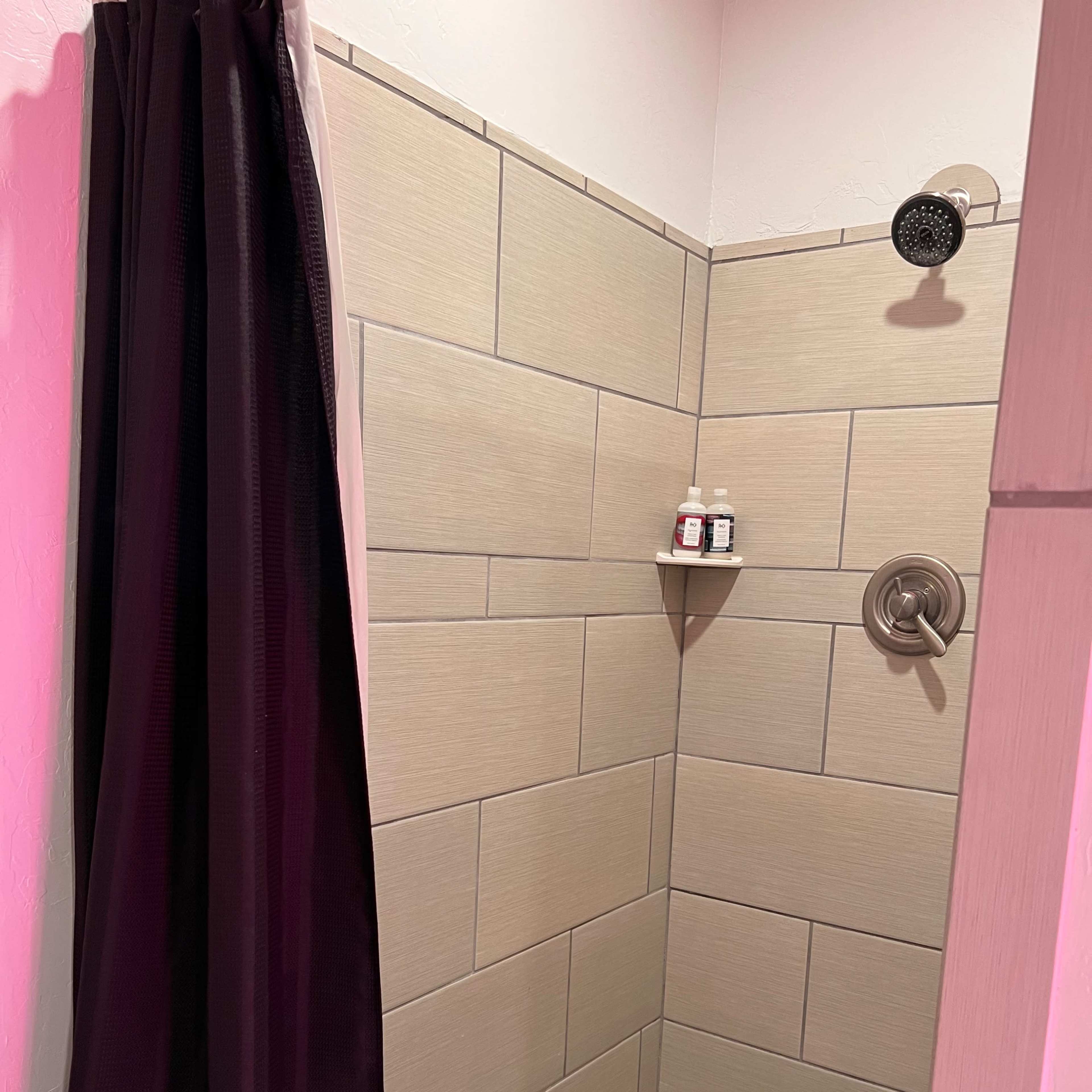 A tiled shower space with a showerhead, a corner shelf containing bottles, and a dark curtain.