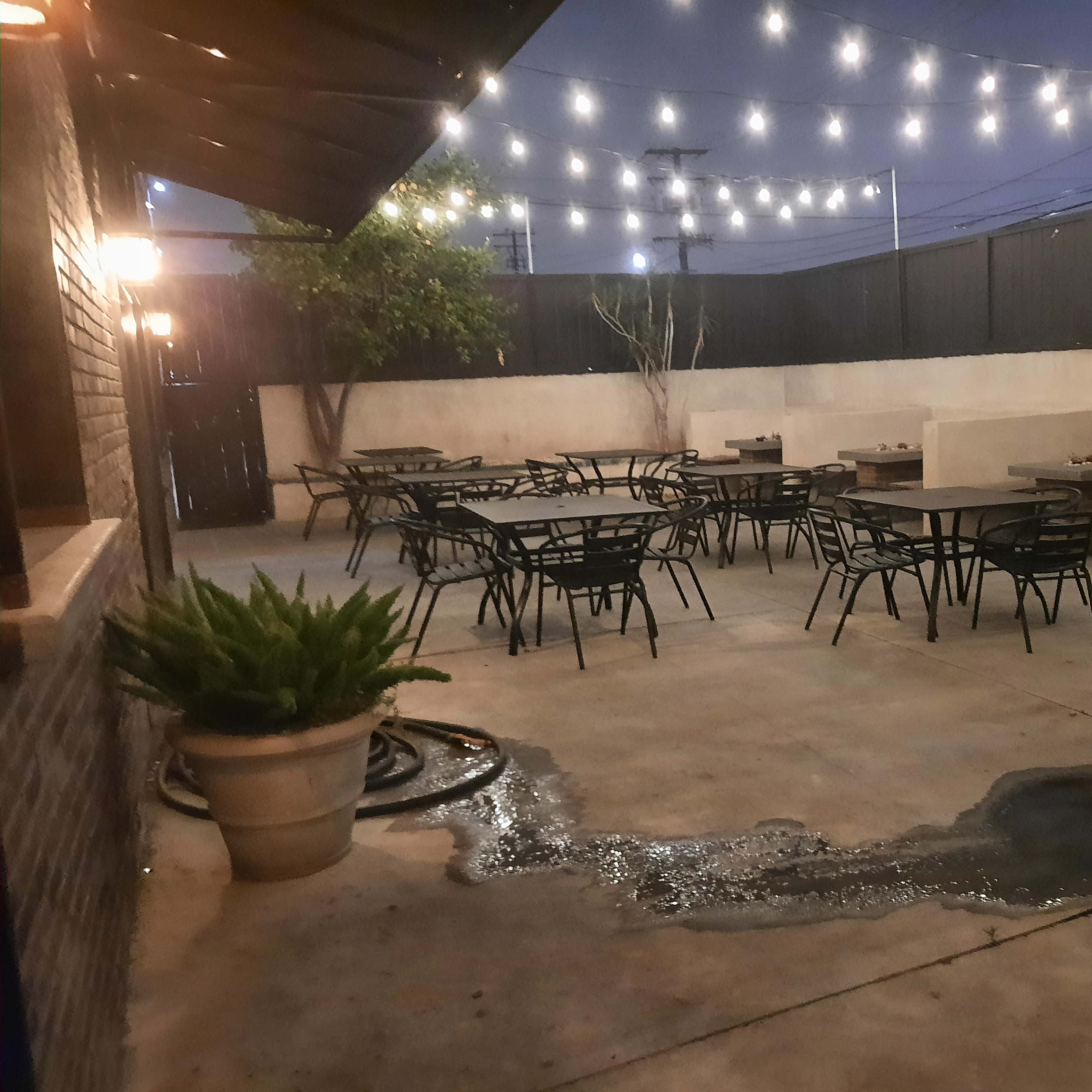 An outdoor dining area features several tables and chairs under hanging lights, with a small puddle of water on the concrete floor.