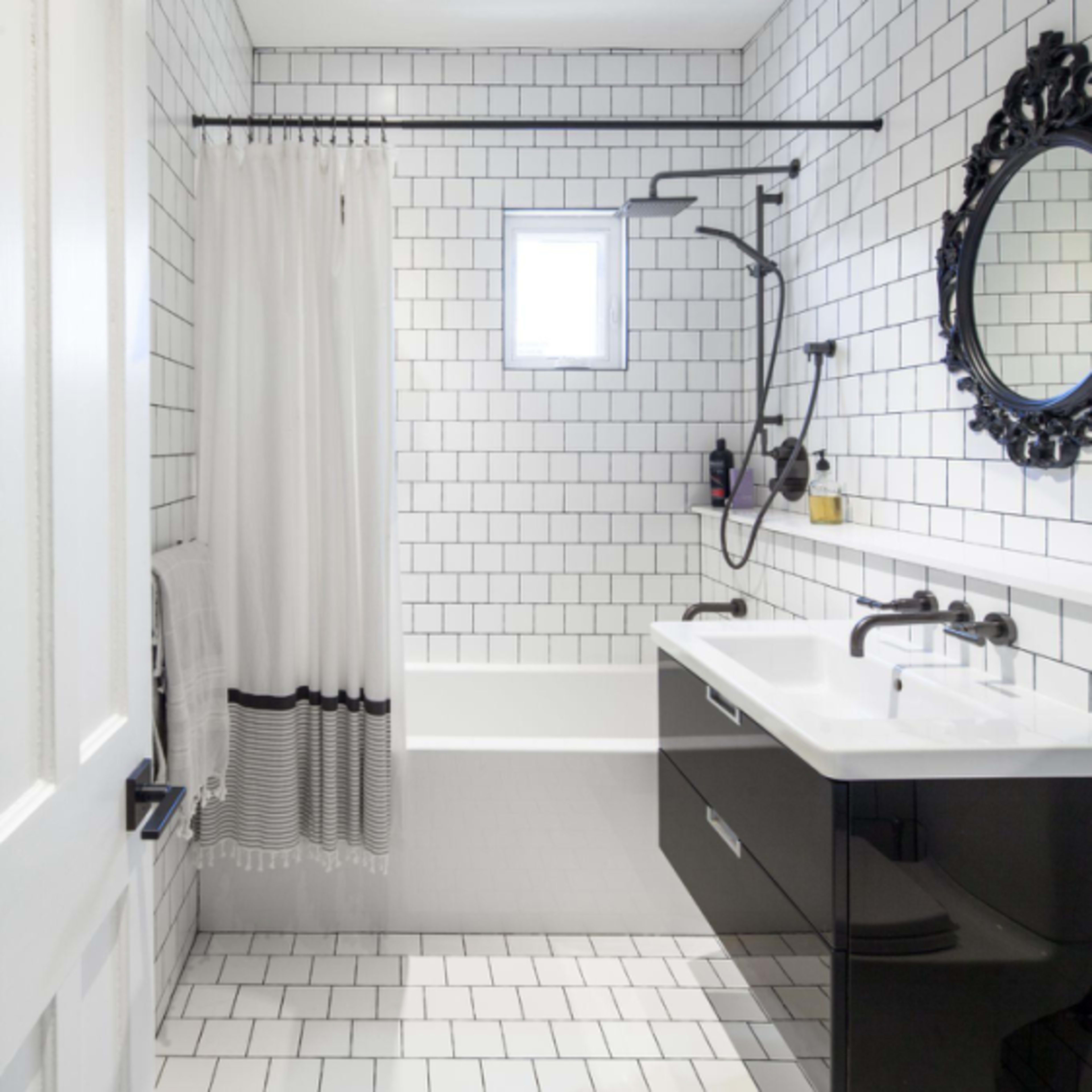 A modern bathroom features white subway tile walls, a black vanity with double sinks, a shower with a glass door, and a window allowing natural light.