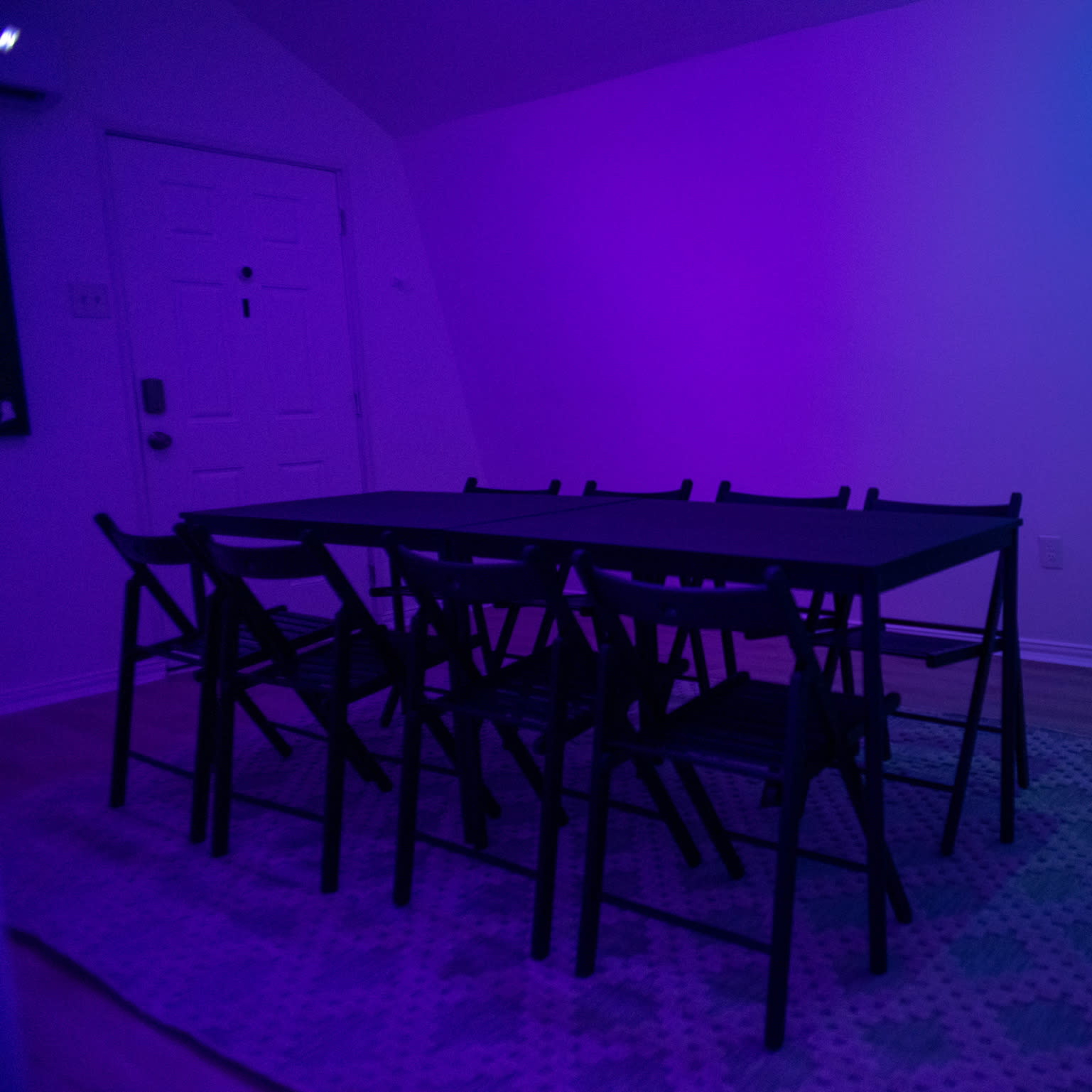A rectangular table with eight black folding chairs sits under purple lighting in a sparsely furnished room, alongside a blank easel.