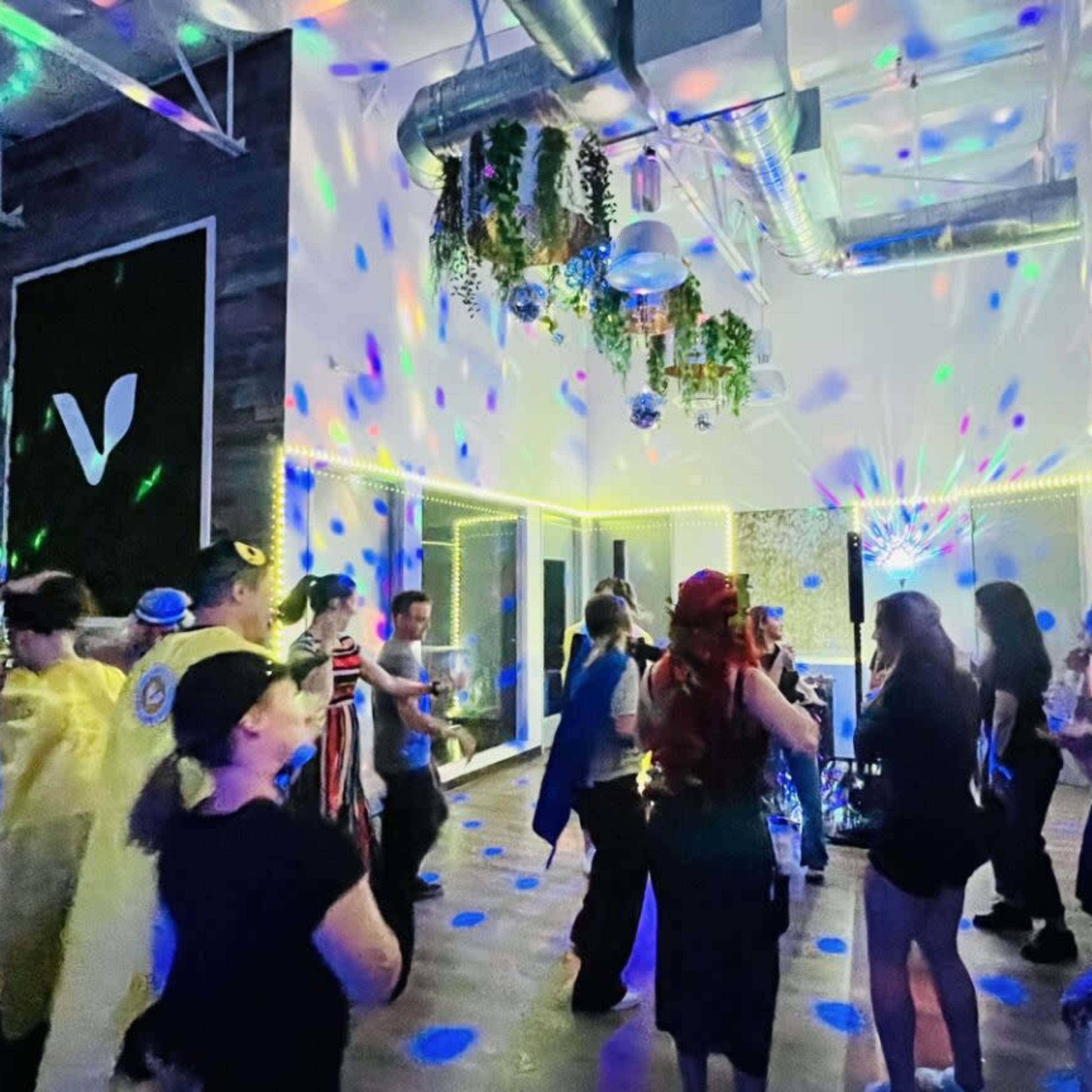 A lively group of people dances under multicolored lights in a spacious indoor venue.