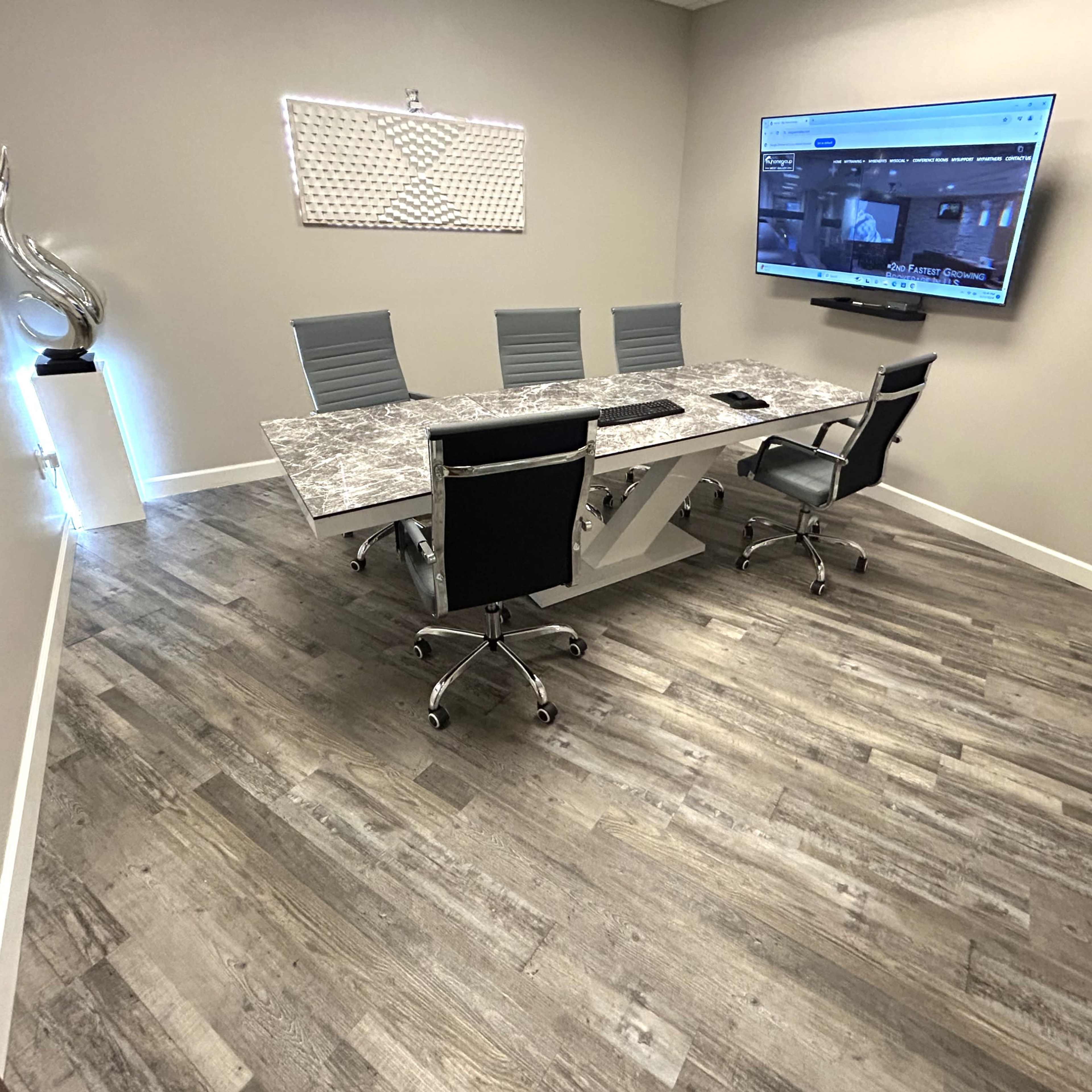A modern conference room with a large table, four chairs, and a wall-mounted screen displaying a video feed.