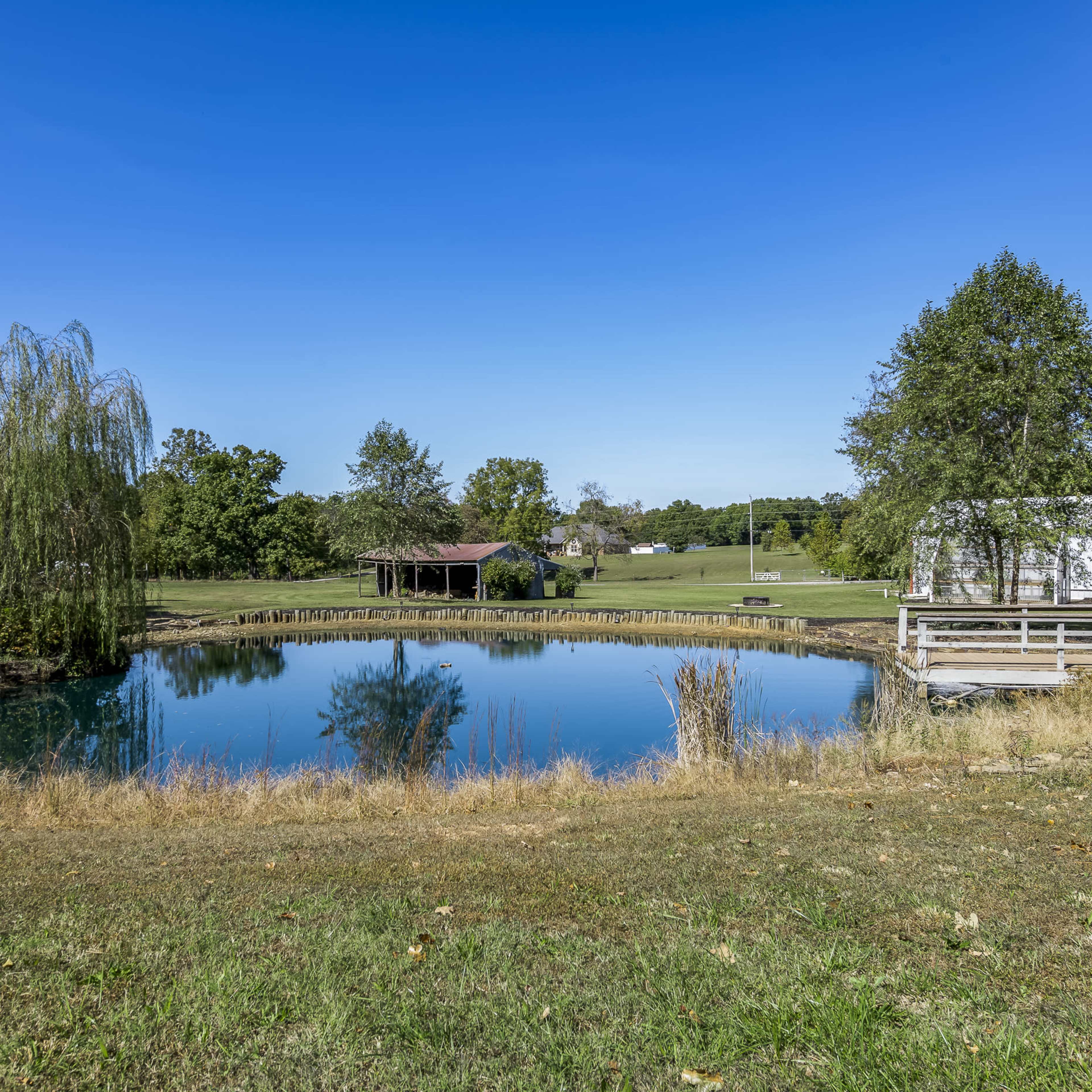 A calm pond surrounded by trees and grassy banks is visible, with a gazebo and white building in the background.