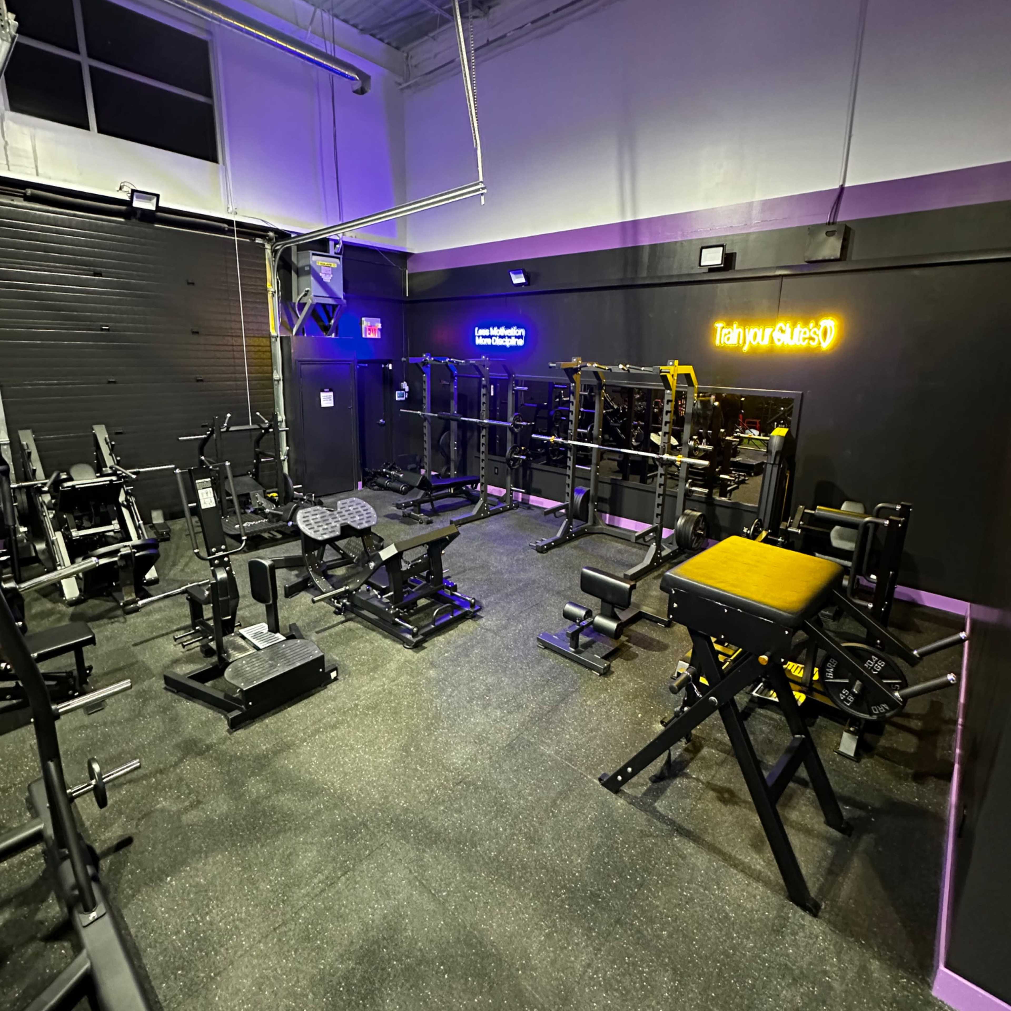 A gym equipped with various weightlifting machines and benches, illuminated by purple lighting.