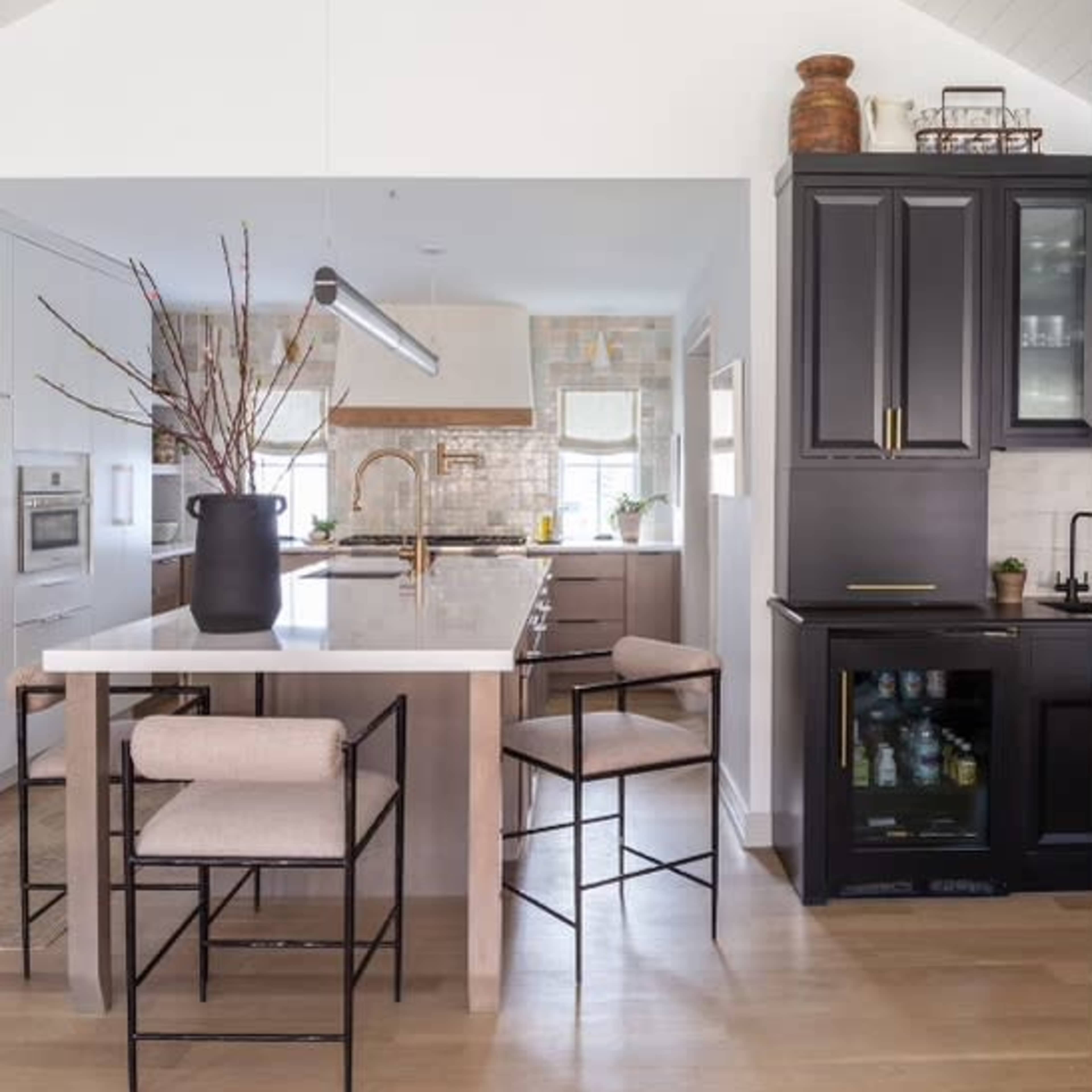 A modern kitchen features a large island with bar stools, a cabinetry section with glass doors, and a high ceiling with exposed beams.