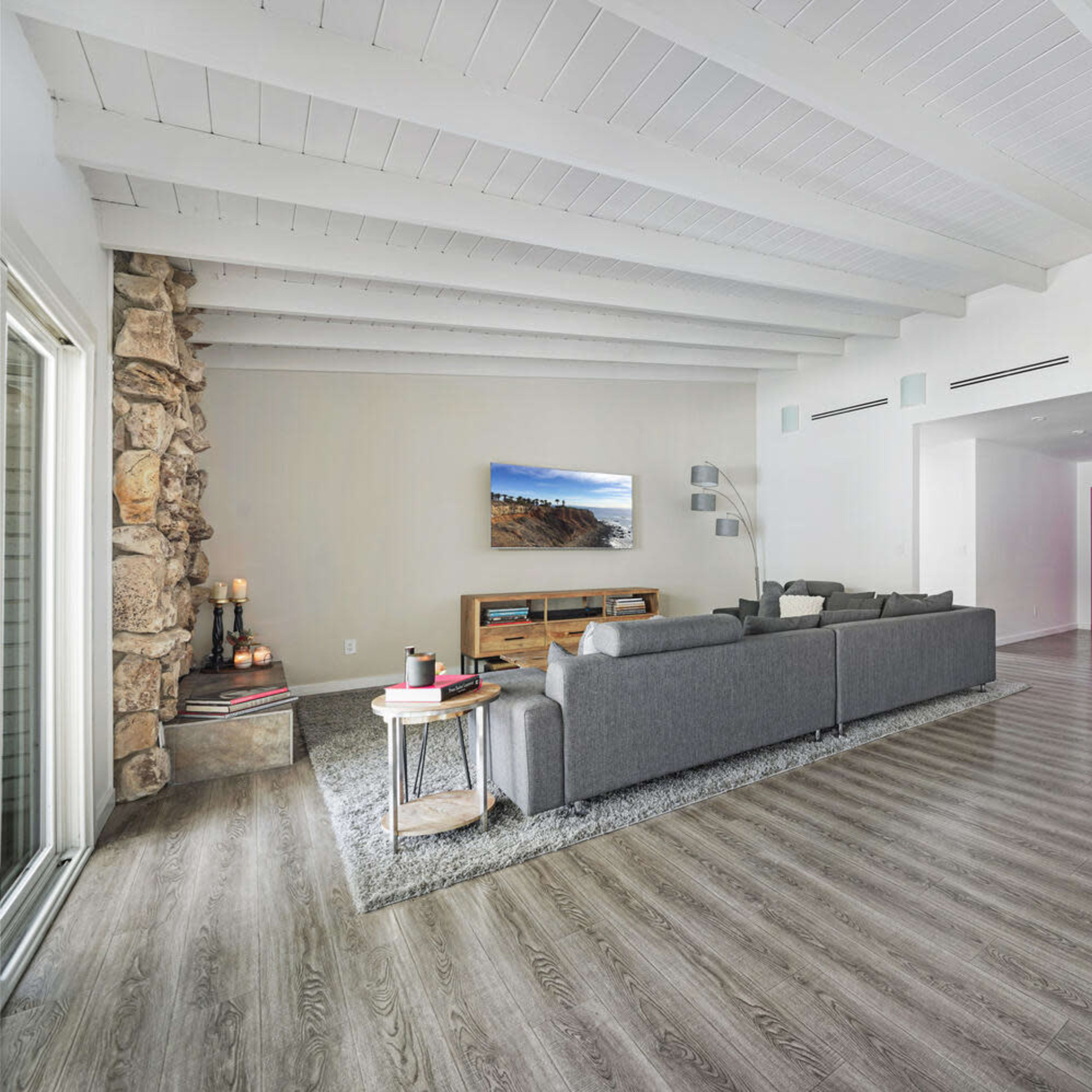 A spacious living area features a gray sectional sofa, wooden media console, and open sightlines toward a modern kitchen with white cabinetry.