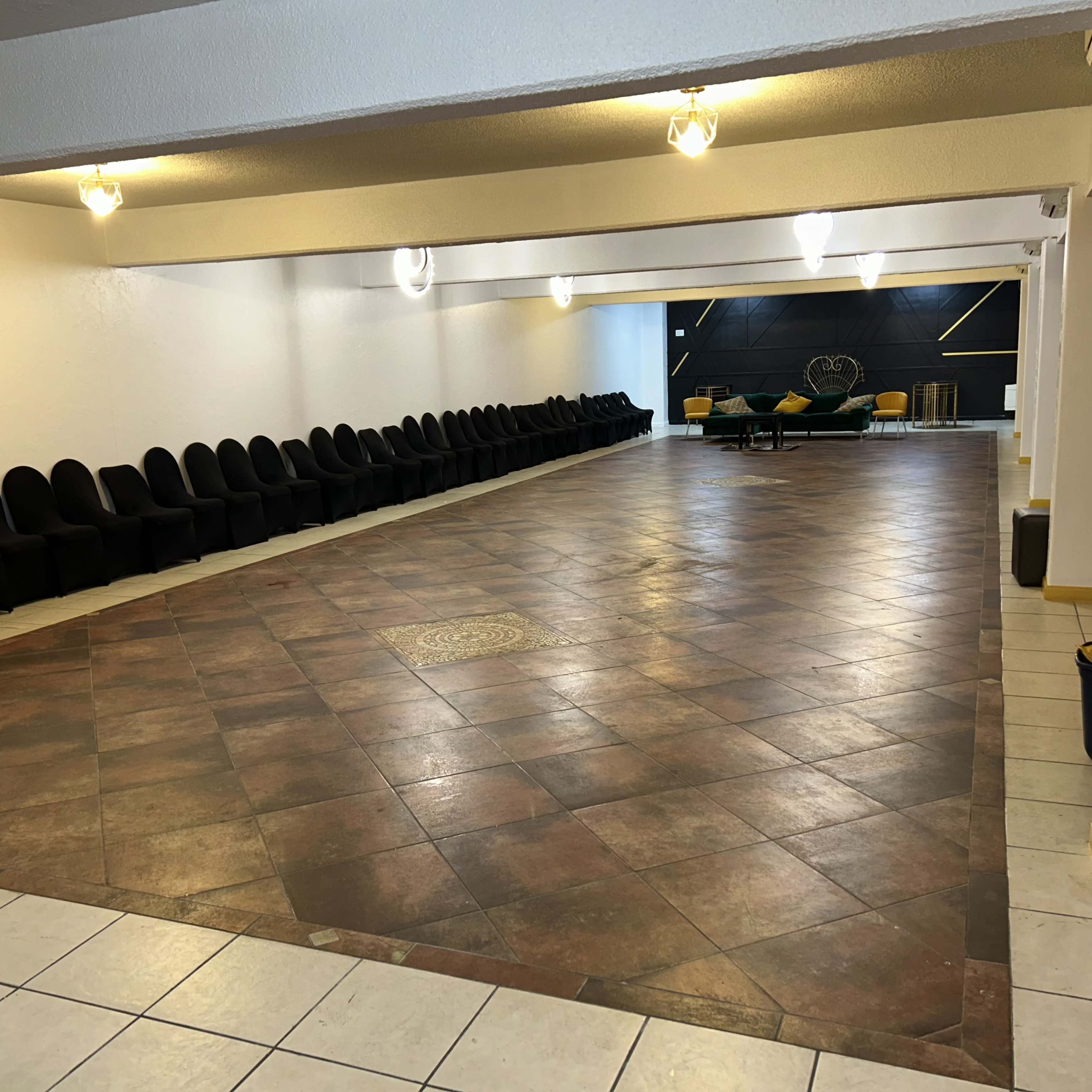 A spacious room with a tiled floor and rows of black chairs along one wall, leading to a seating area with green and yellow furniture at the far end.
