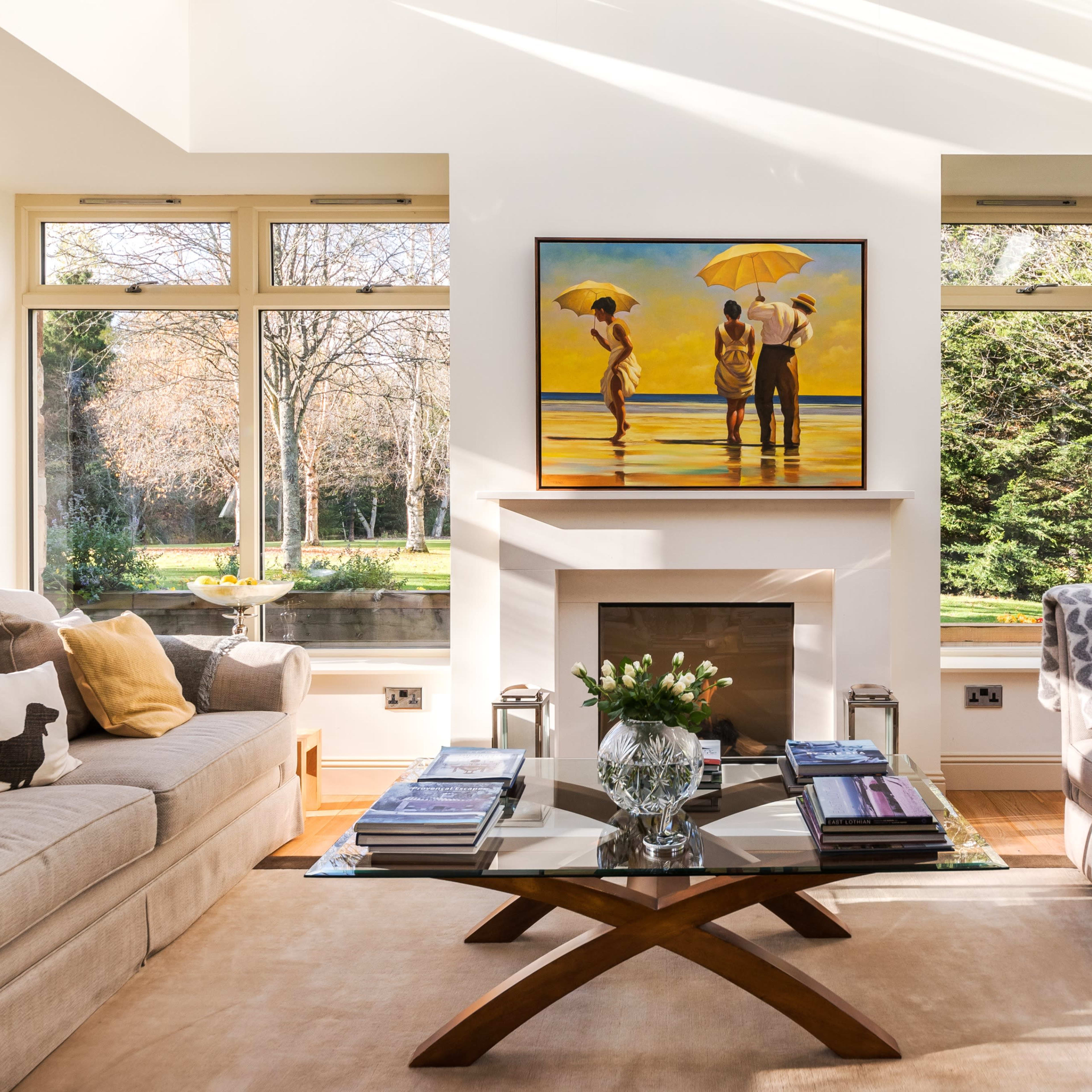 The living room features two sofas facing a glass coffee table, with a painting of figures holding umbrellas above a fireplace and large windows displaying a view of trees outside.