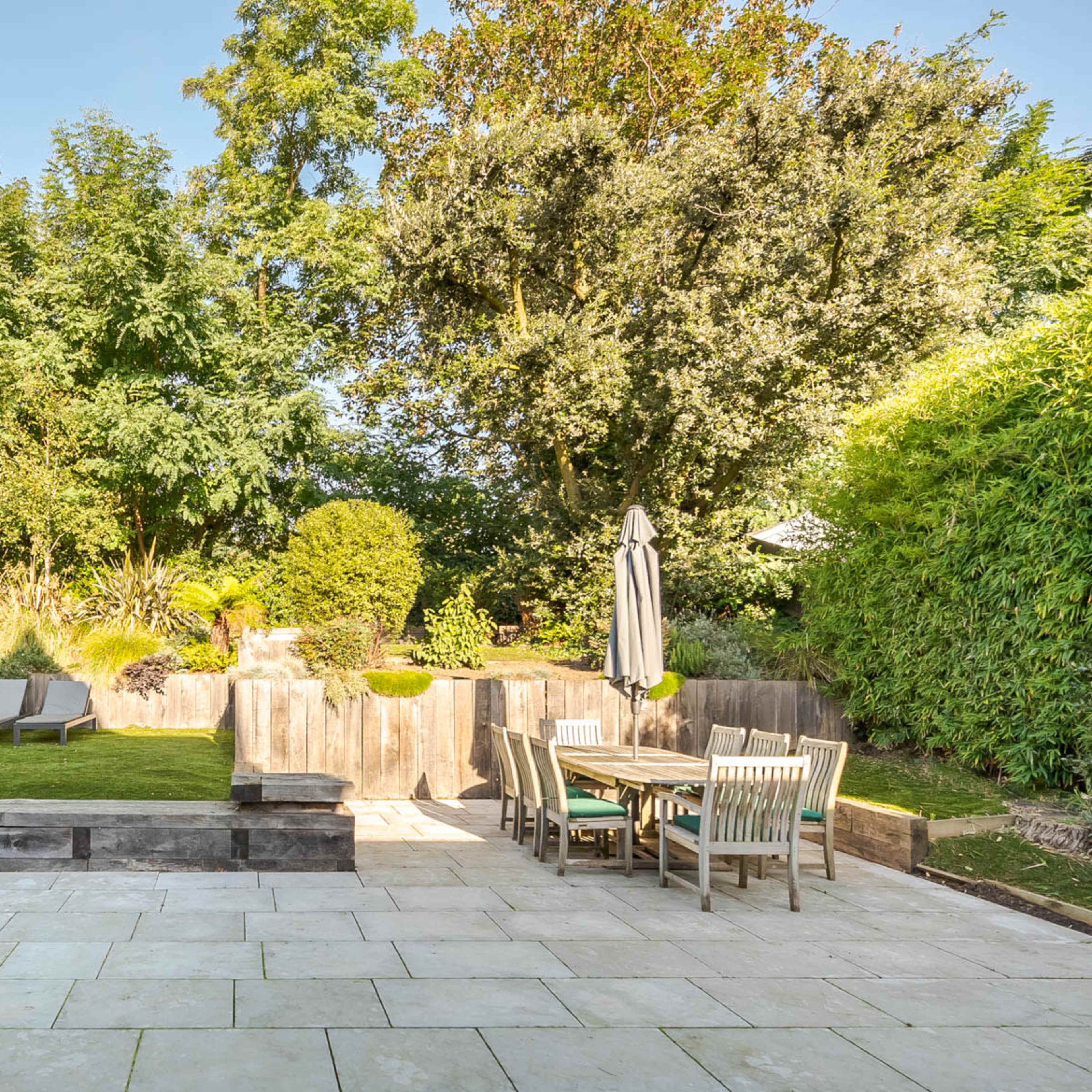 A landscaped garden featuring a grassy area, wooden seating, and a dining table surrounded by tall trees and hedges.