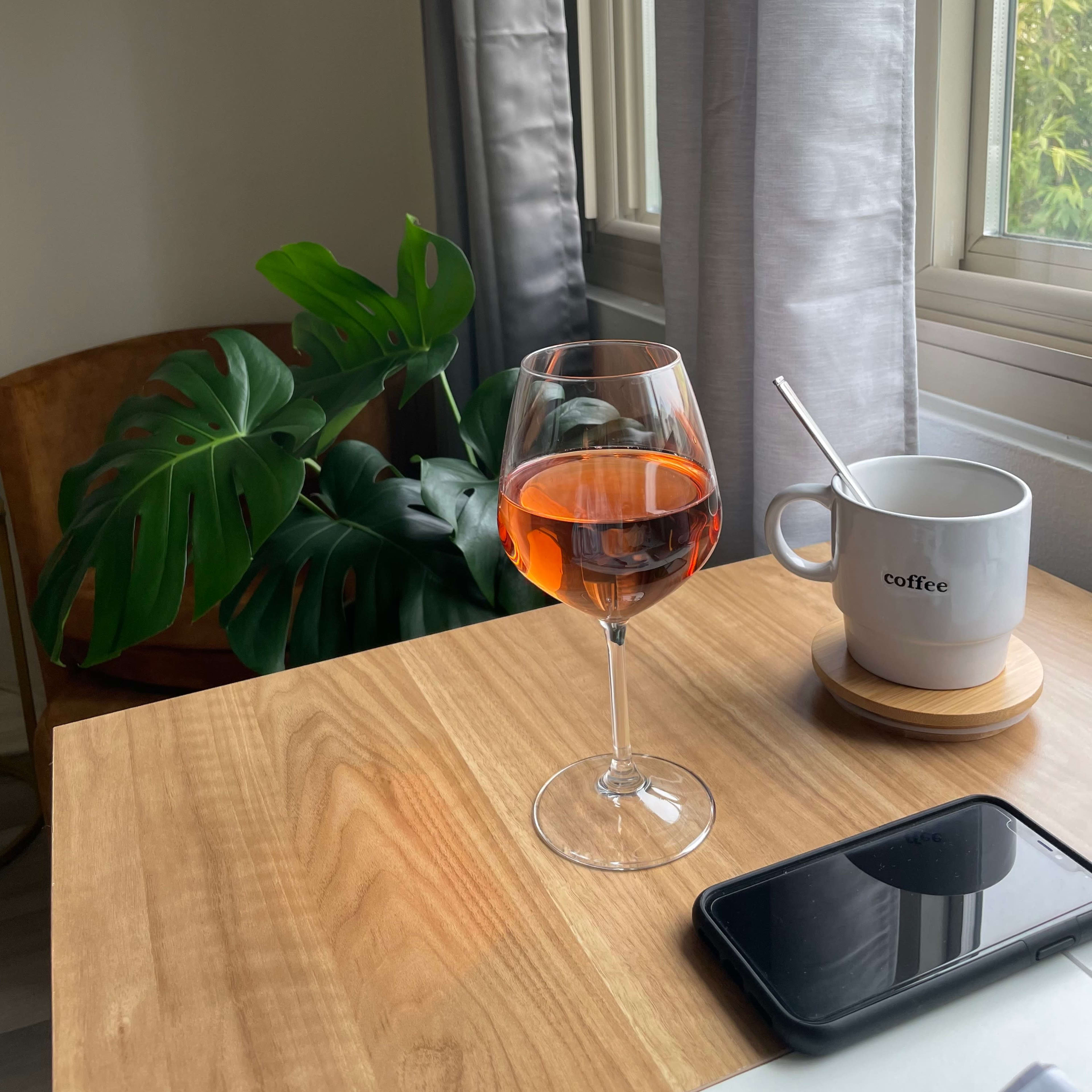 A glass of rosé wine sits on a wooden table next to a coffee cup and a smartphone, with a large green plant in the background.