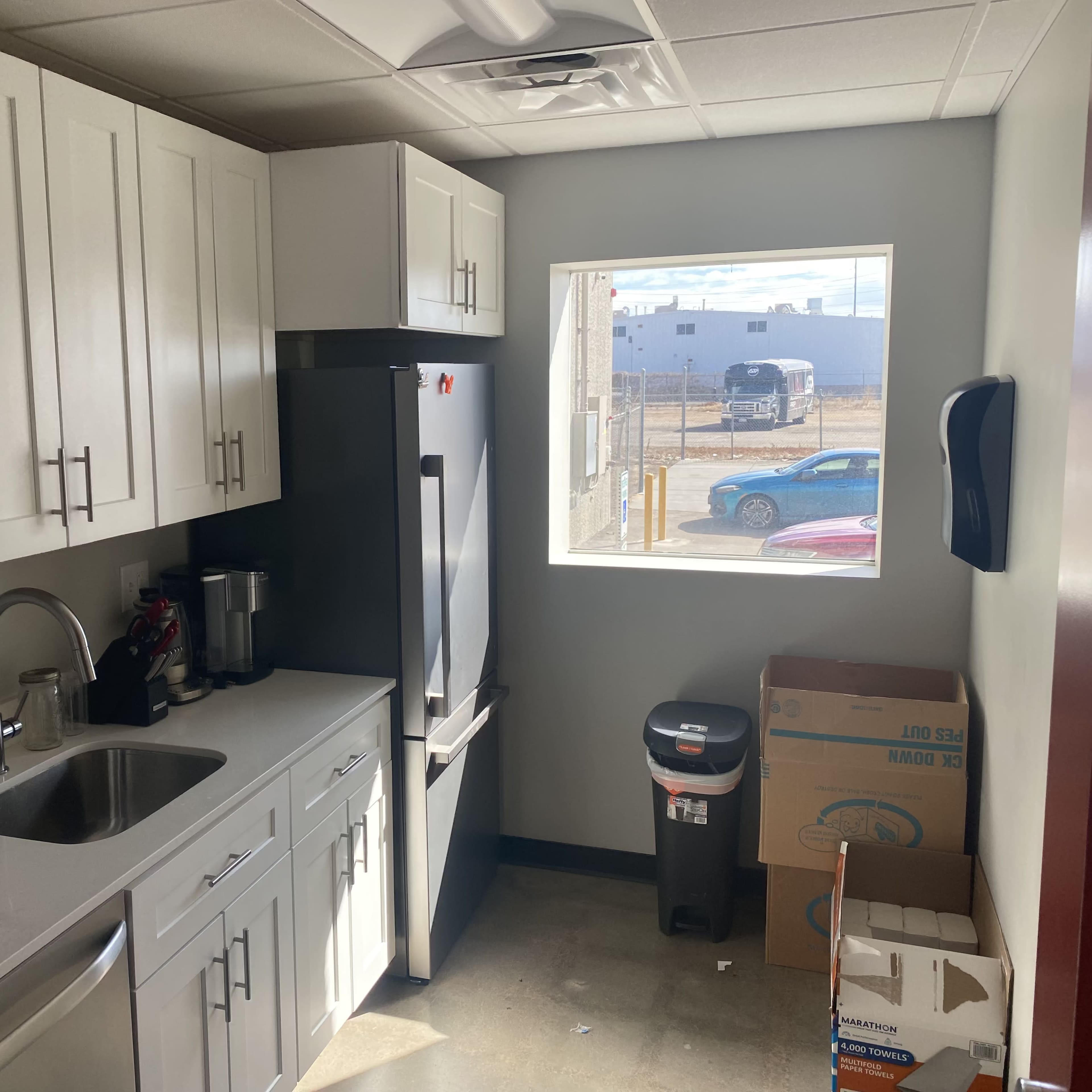 A kitchen with white cabinets, a stainless steel refrigerator, a sink, and a window showing an outdoor scene, along with a trash can and cardboard boxes.