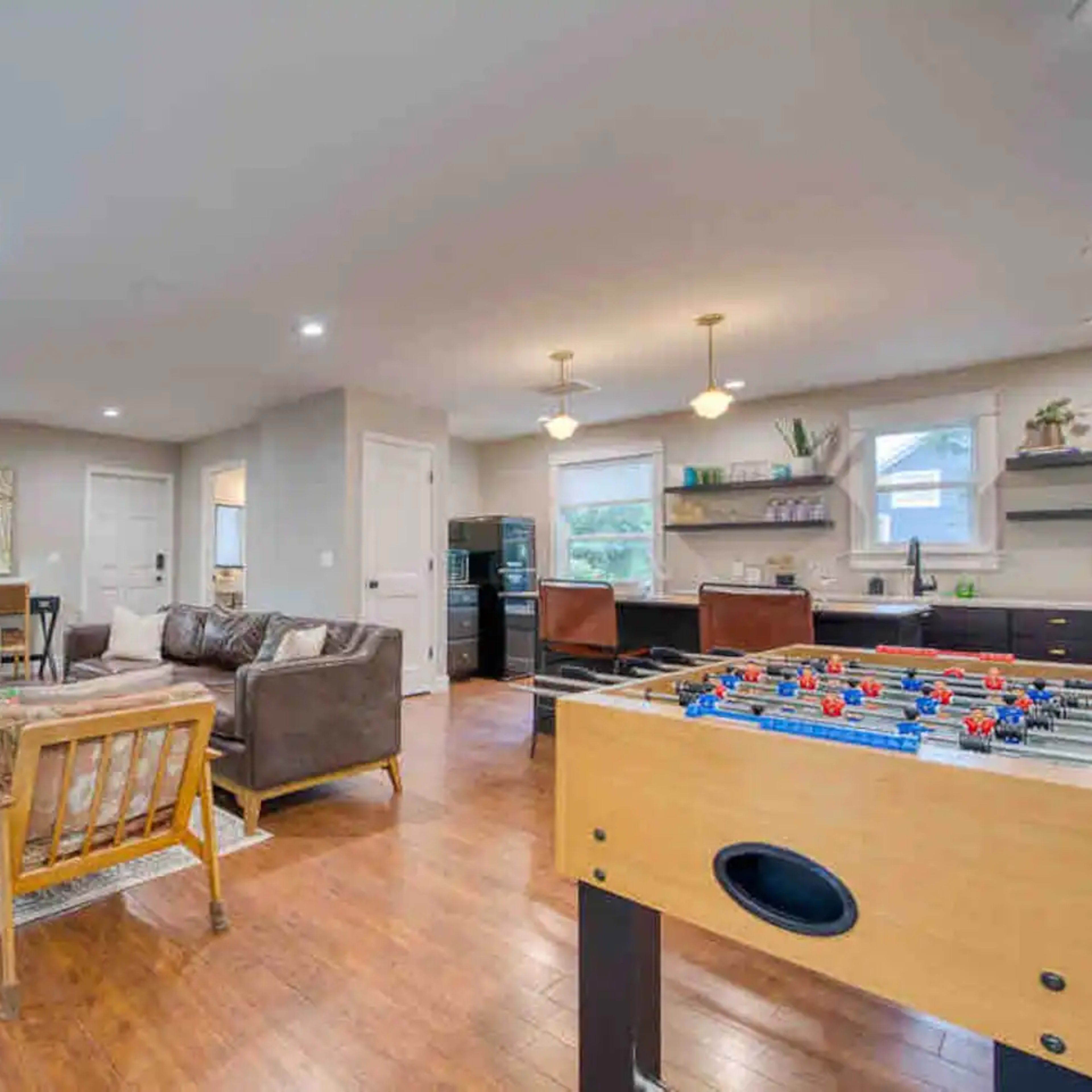 A modern living space featuring a foosball table, a seating area with two sofas, a coffee table, and a kitchen in the background.