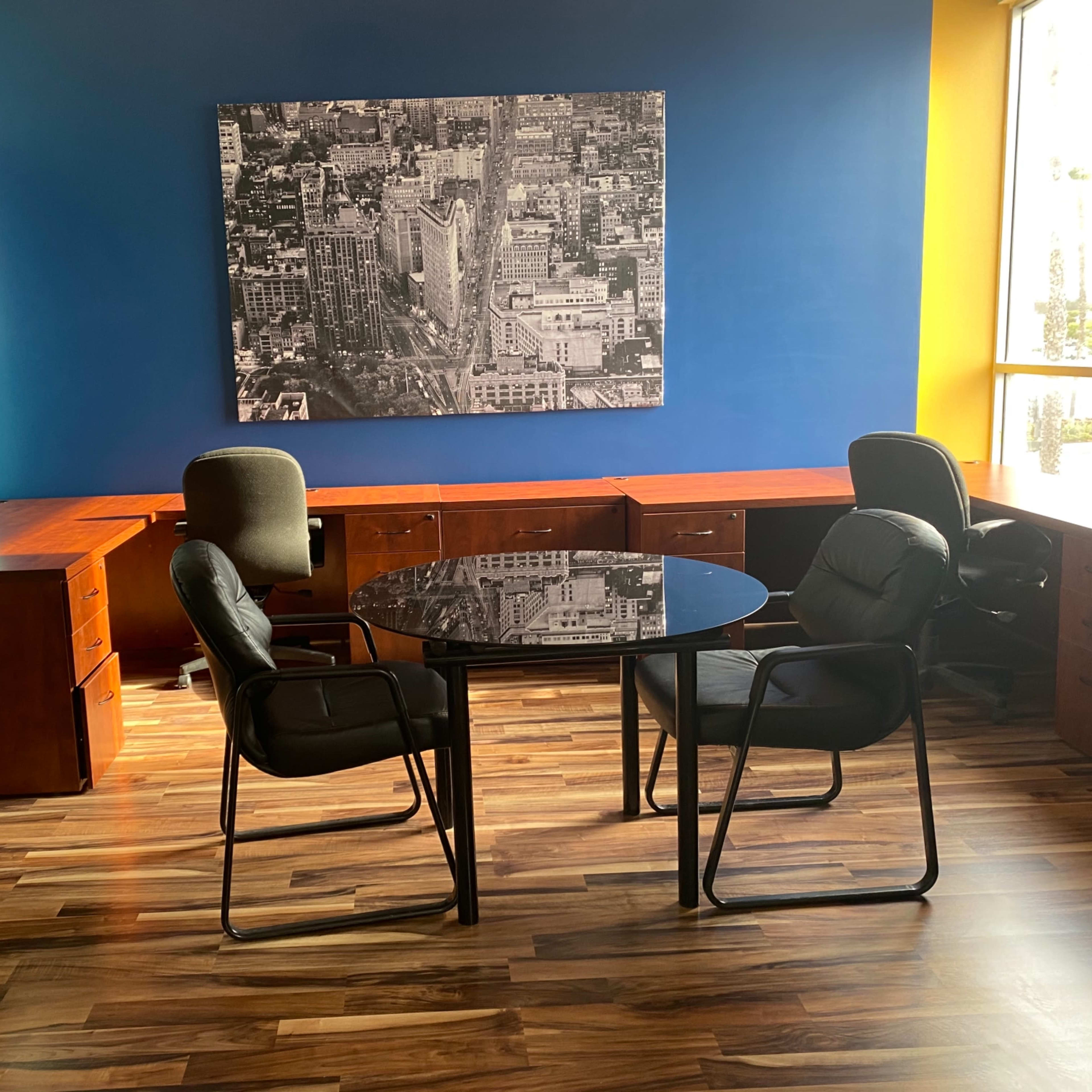 An office space with a round black table and two chairs, surrounded by wooden desks and a large black-and-white photograph on the wall.
