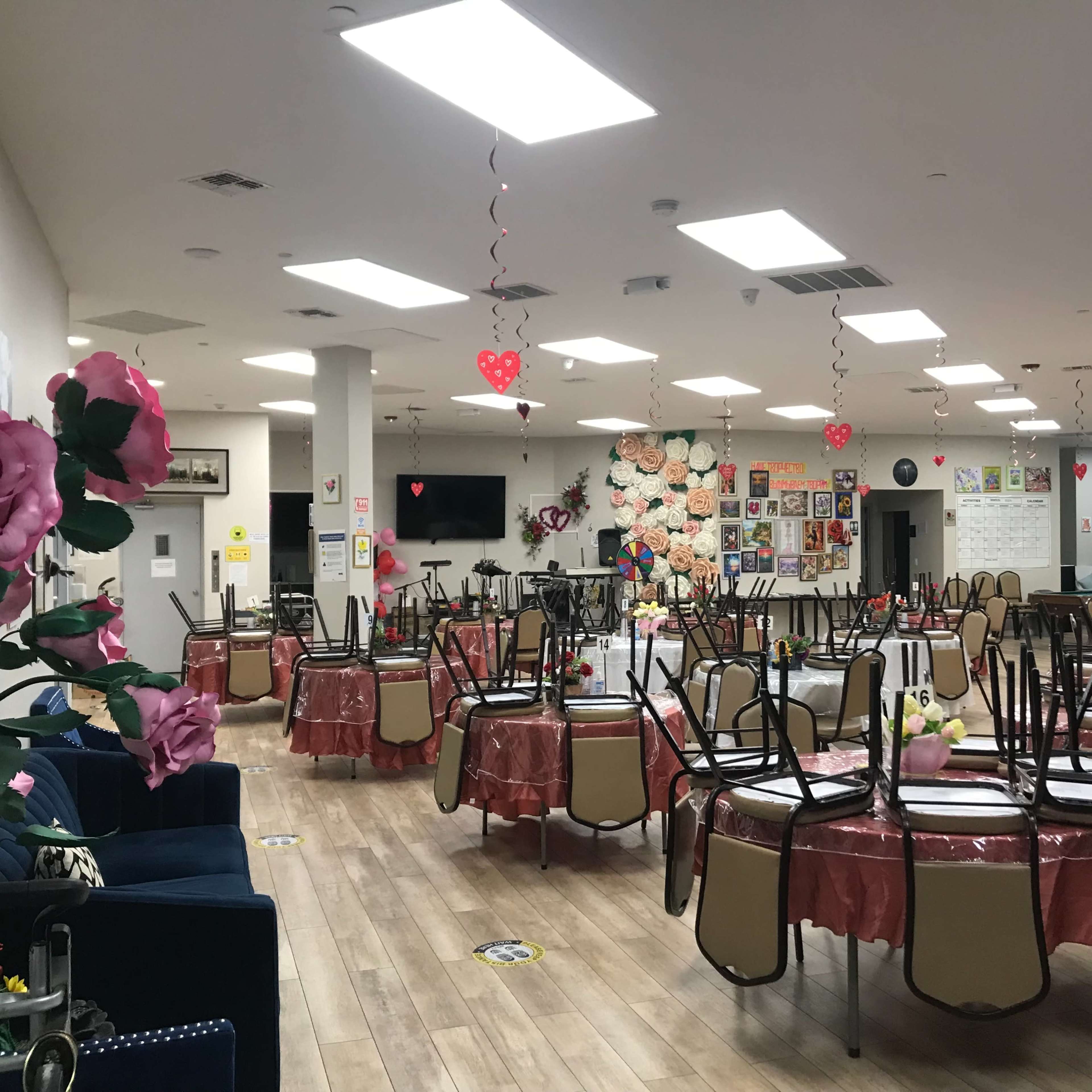 A large, decorated dining area with tables set for a gathering, featuring floral centerpieces and heart-shaped decorations hanging from the ceiling.