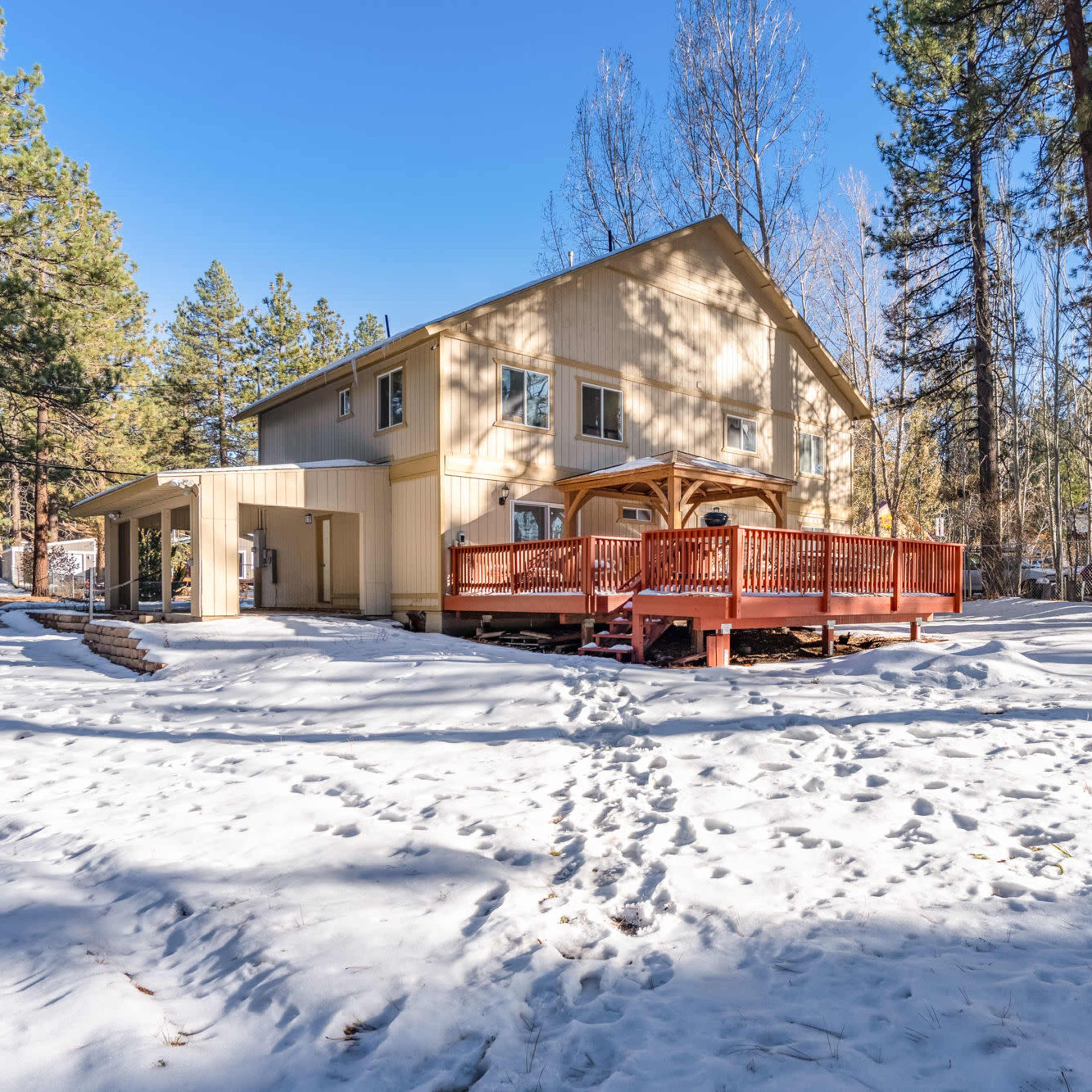A two-story house with a red deck stands among snow-covered ground and tall pine trees.