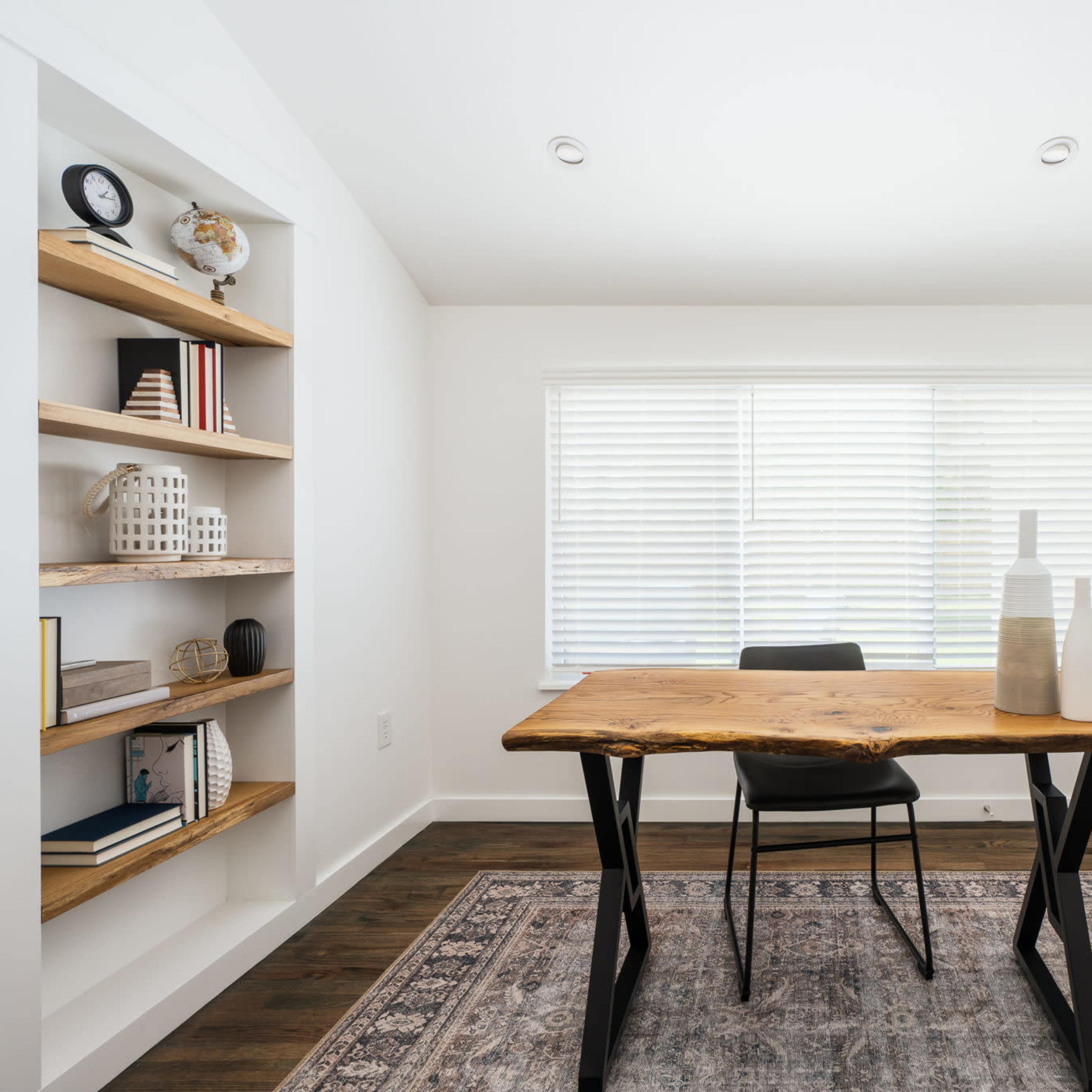 A minimalist home office features a wooden desk with a black chair, a shelf filled with books and decorative items, and a window covered with blinds.