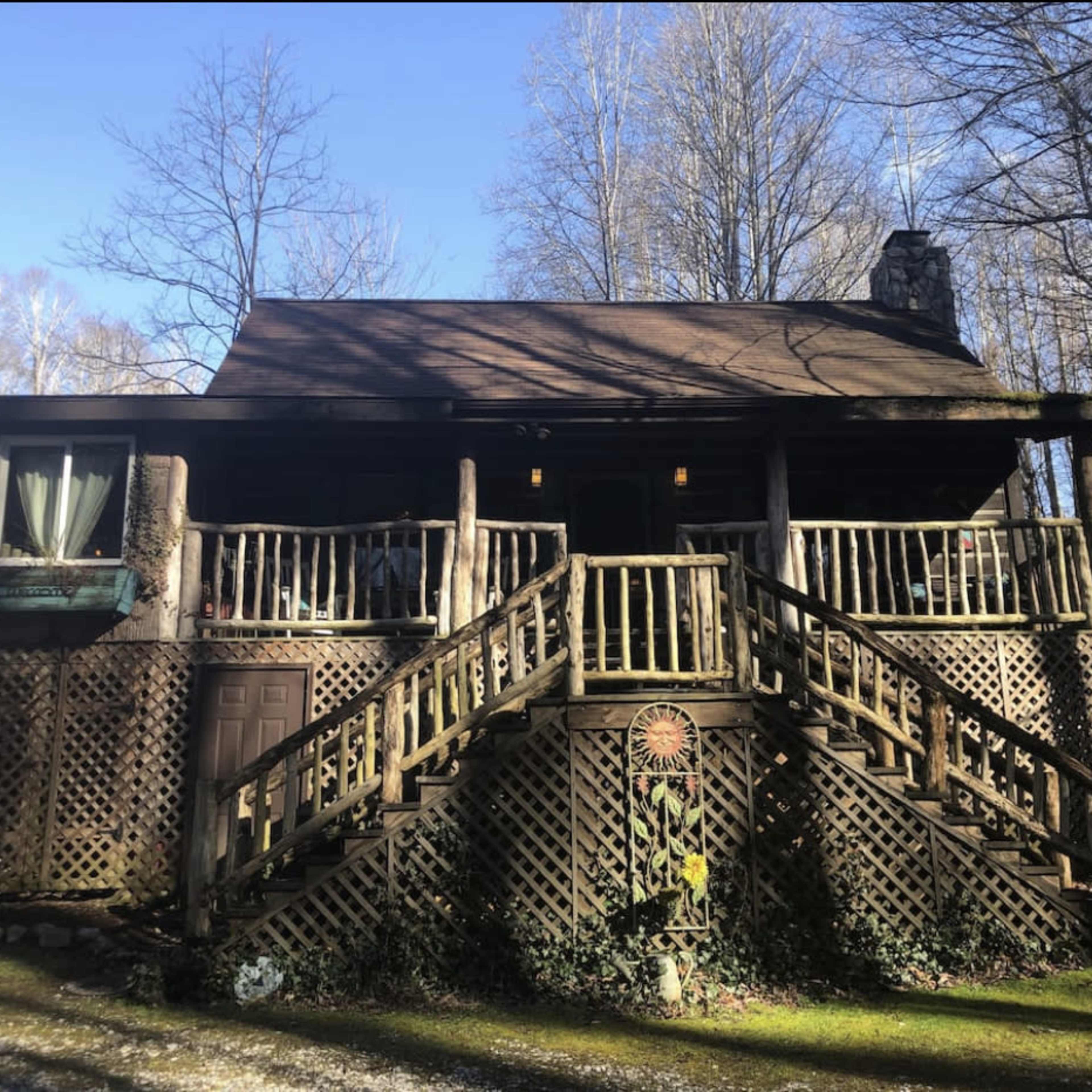 A rustic cabin with wooden lattice and two sets of stairs is surrounded by trees in a forested area.