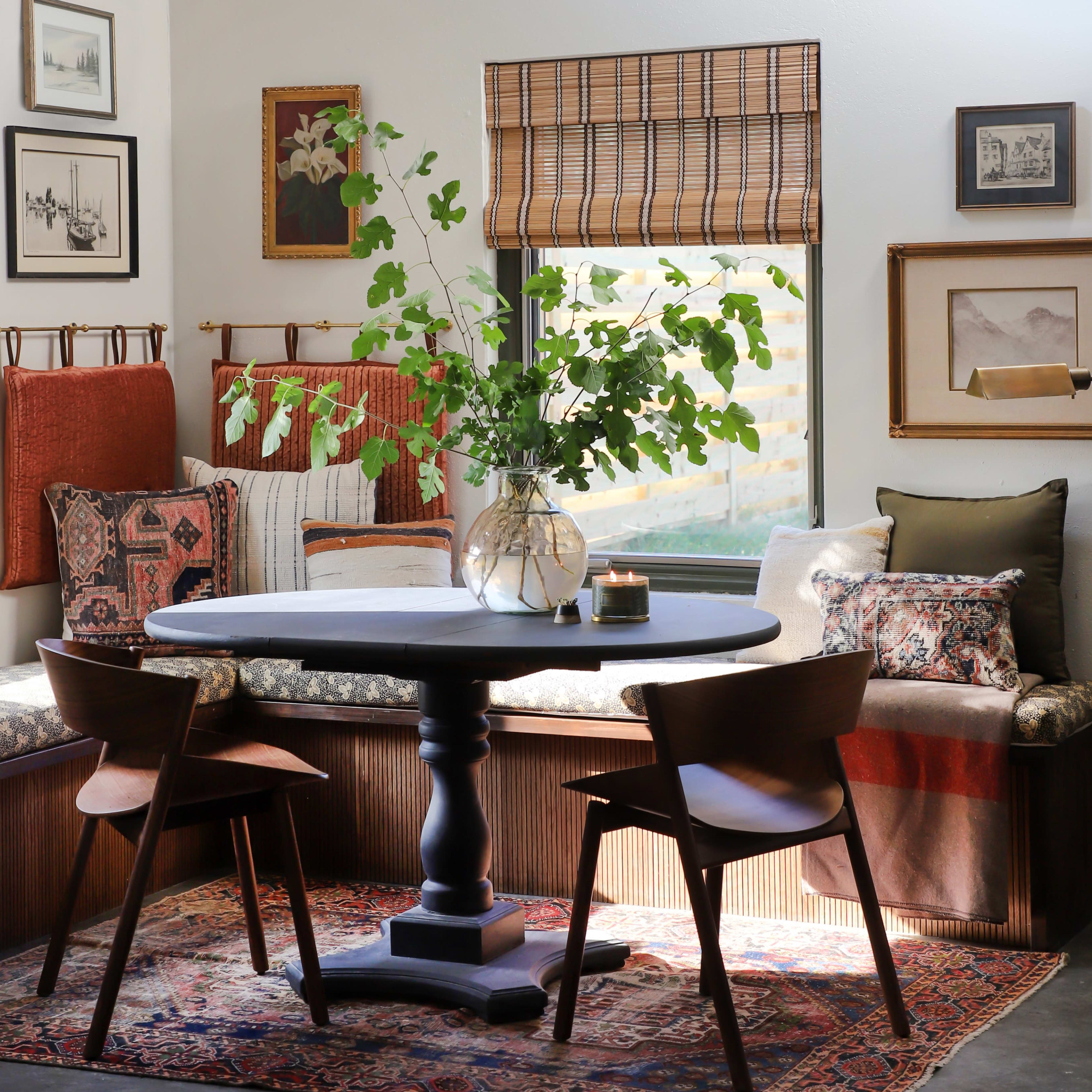 A cozy dining area featuring a round table with two chairs, surrounded by a window seat adorned with patterned pillows and framed artwork on the walls.