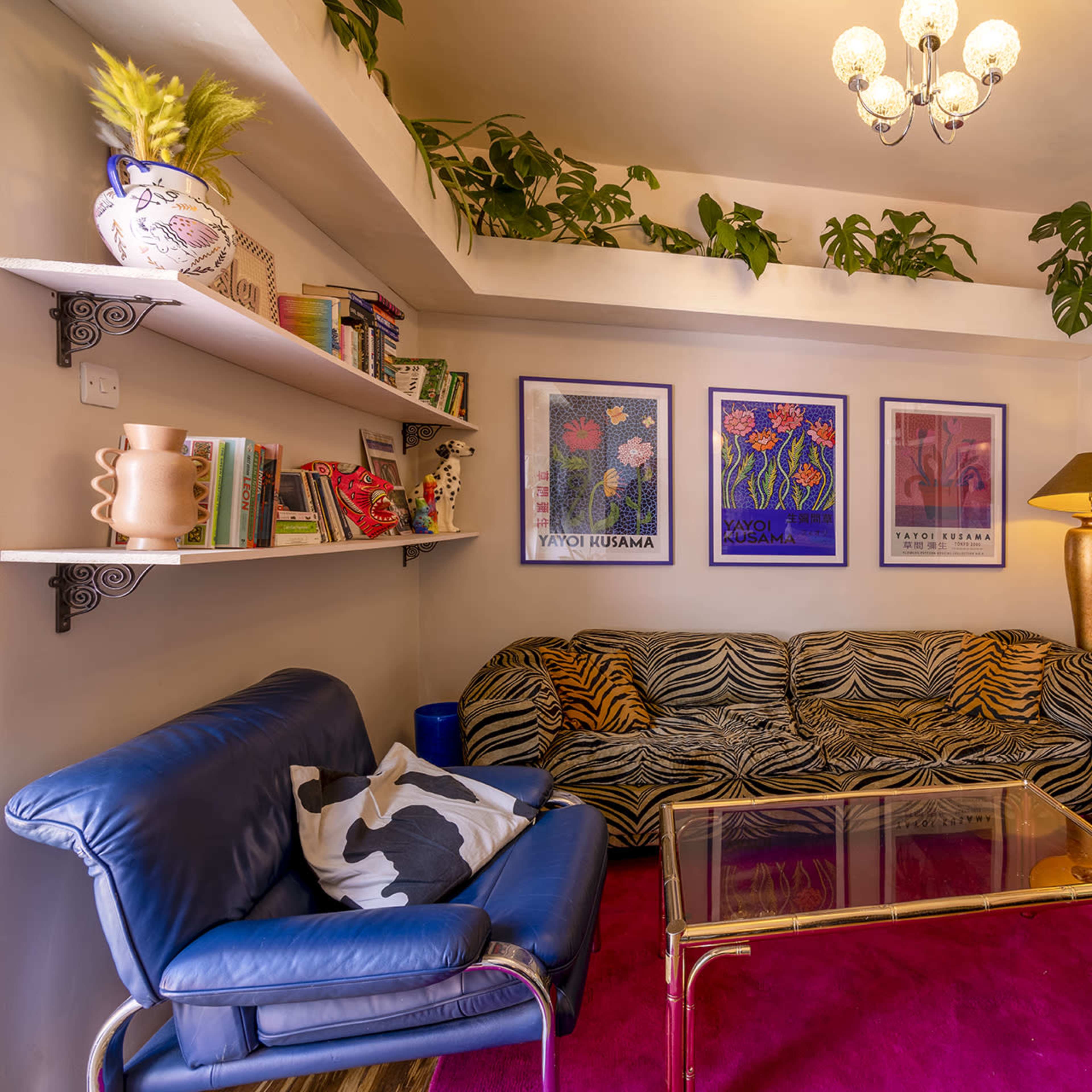 A living room with a zebra-striped couch, blue chairs, a glass coffee table, colorful wall art, potted plants, and a light fixture.