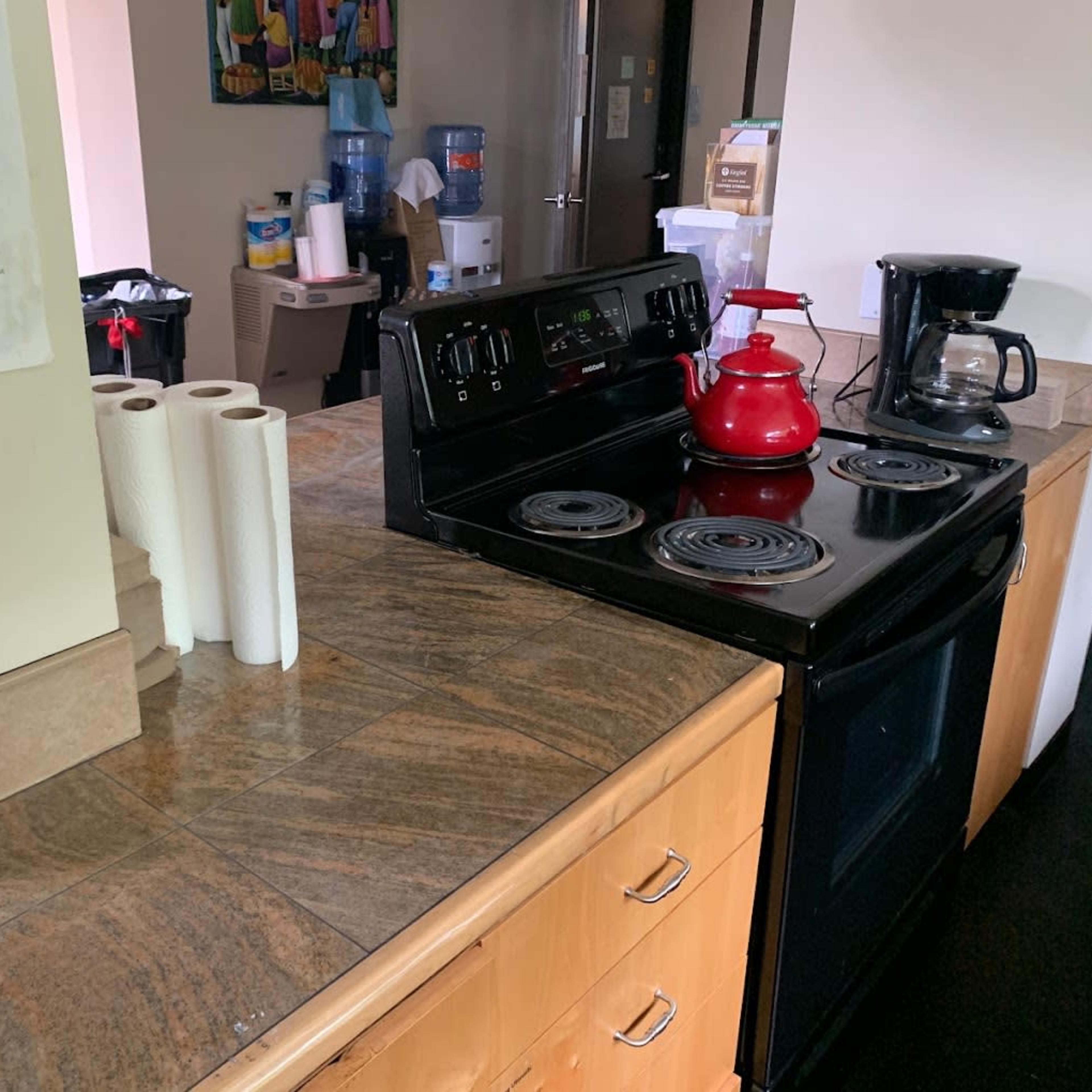 A kitchen counter with a black stove, a red kettle, a coffee maker, paper towel rolls, and a water cooler in the background.