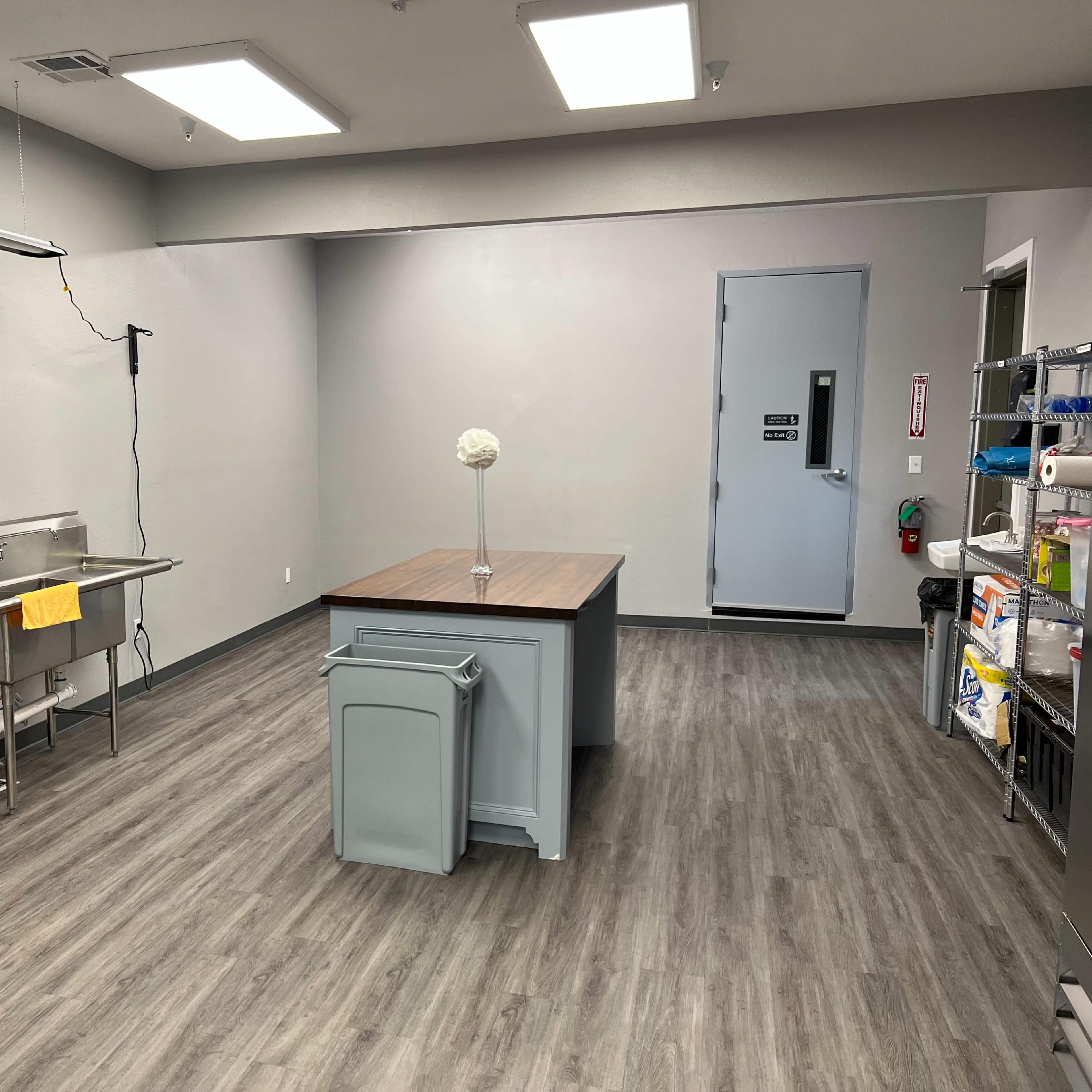 A brightly lit room with gray walls featuring a stainless steel sink on one side, a central table with a wooden top, and a door at the back.