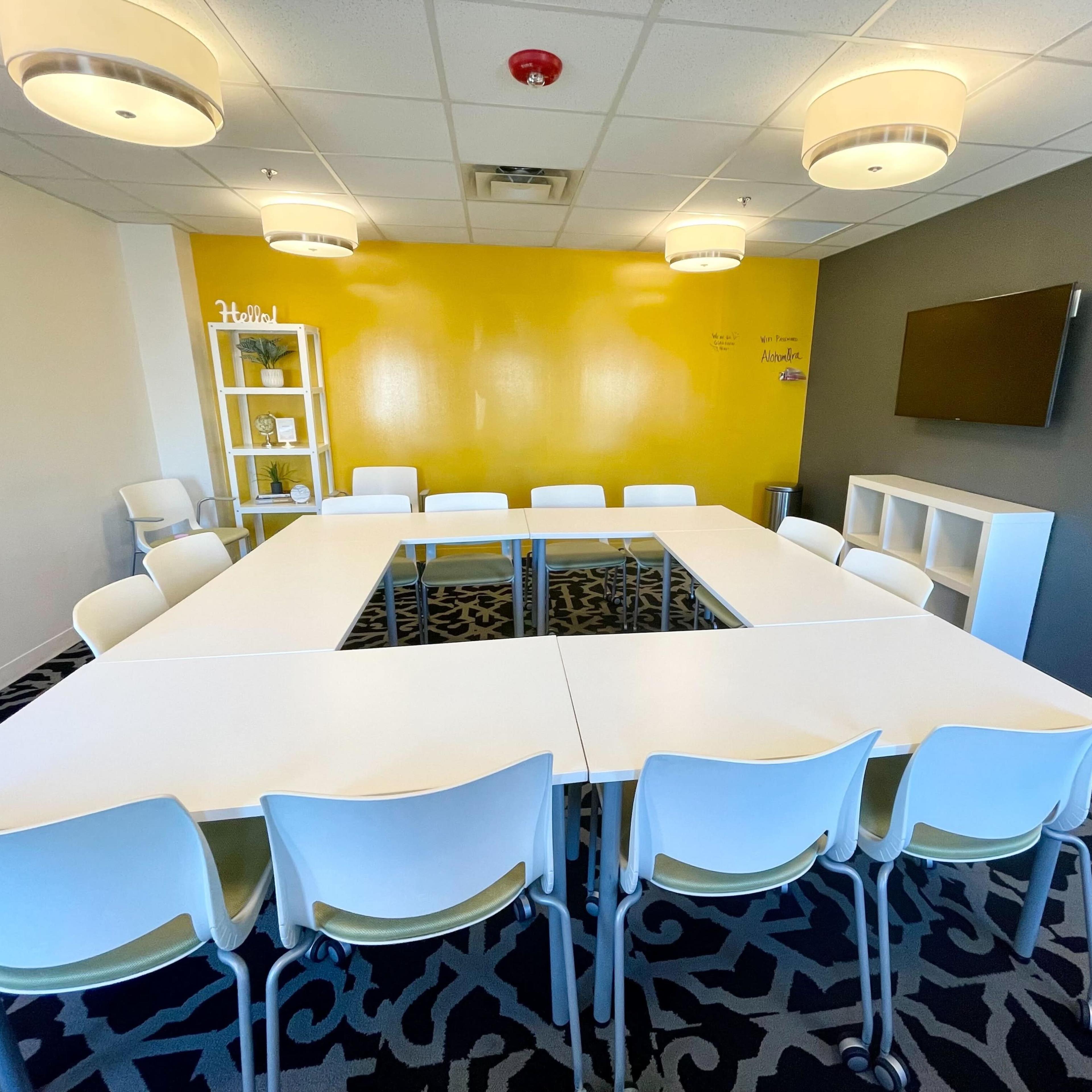 A modern conference room featuring a large rectangular table surrounded by white chairs, with a yellow accent wall and a television mounted on the gray wall.