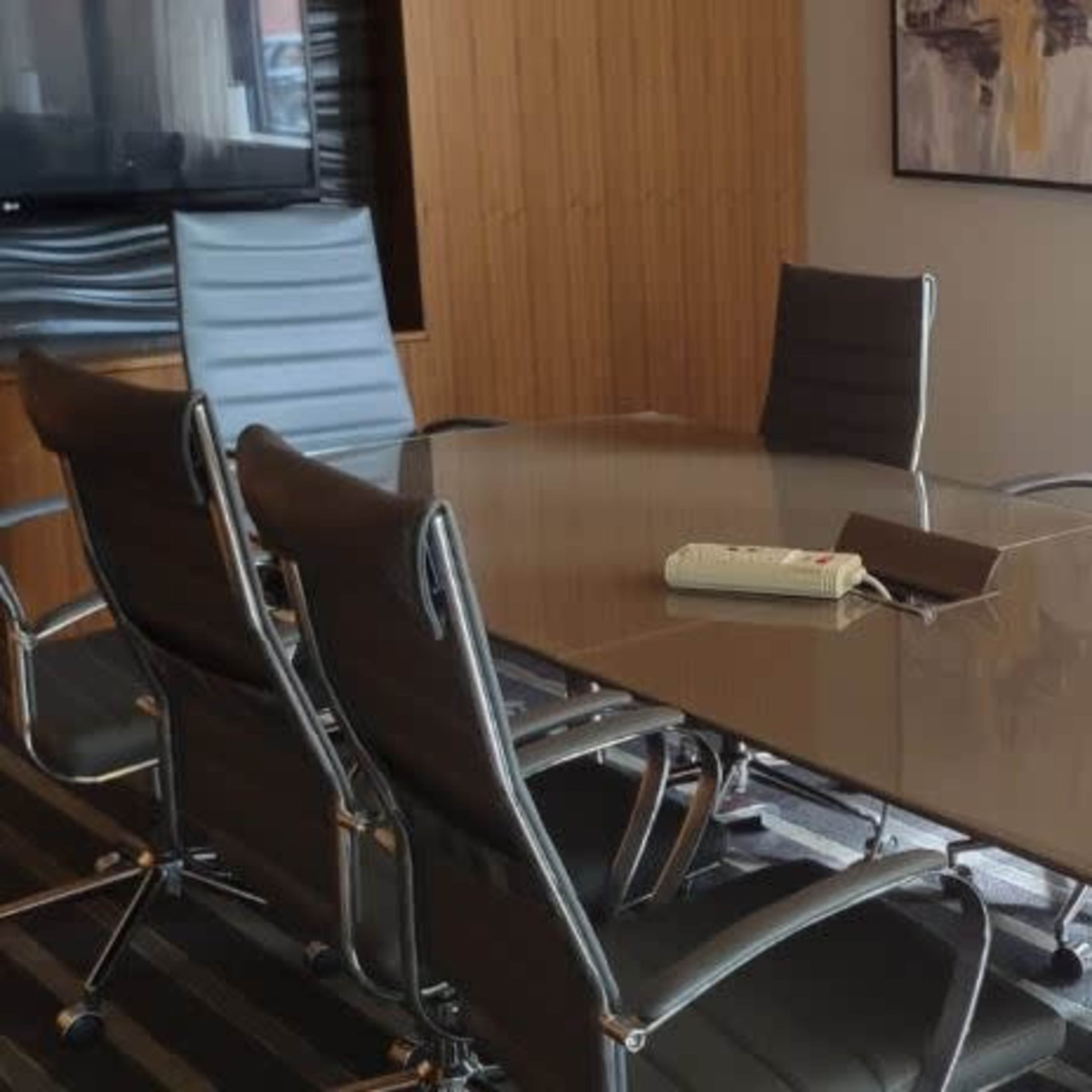 A modern conference room features a large glass table surrounded by eight sleek chairs, with natural light streaming through a nearby window.