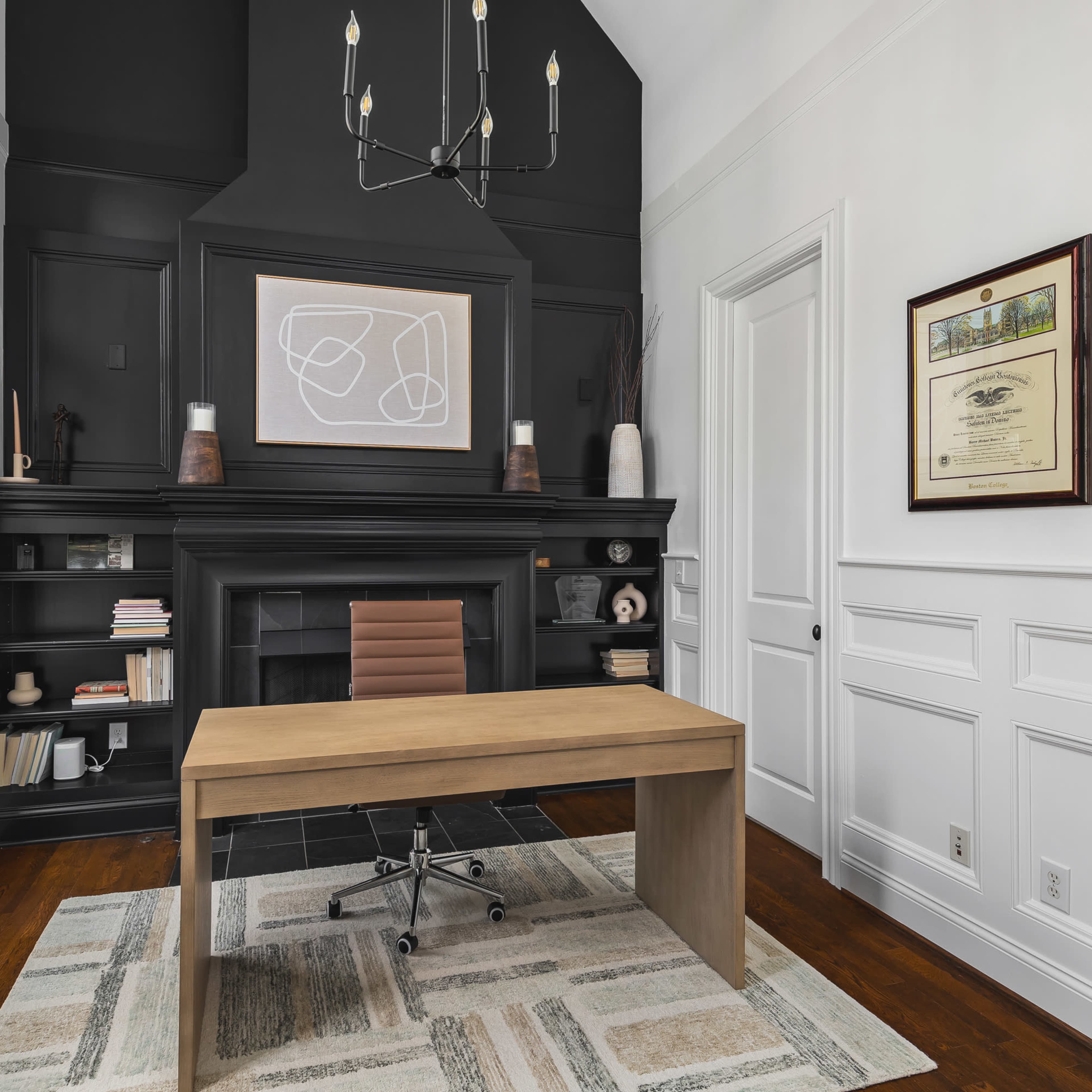 A modern office with a wooden desk facing a large black fireplace, surrounded by white walls and tall windows that let in natural light.