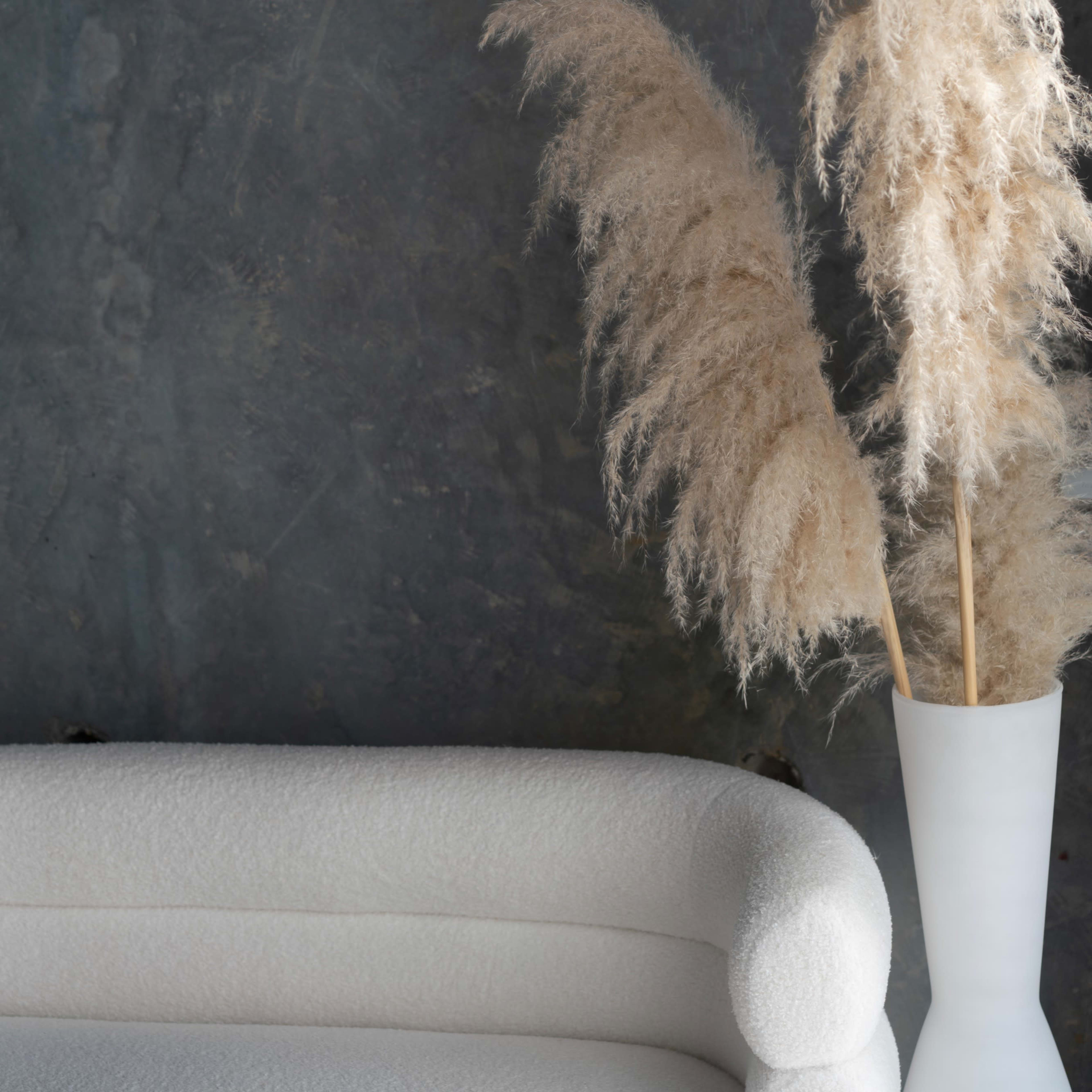 A white couch is positioned next to a tall vase containing dried pampas grass against a dark textured wall.