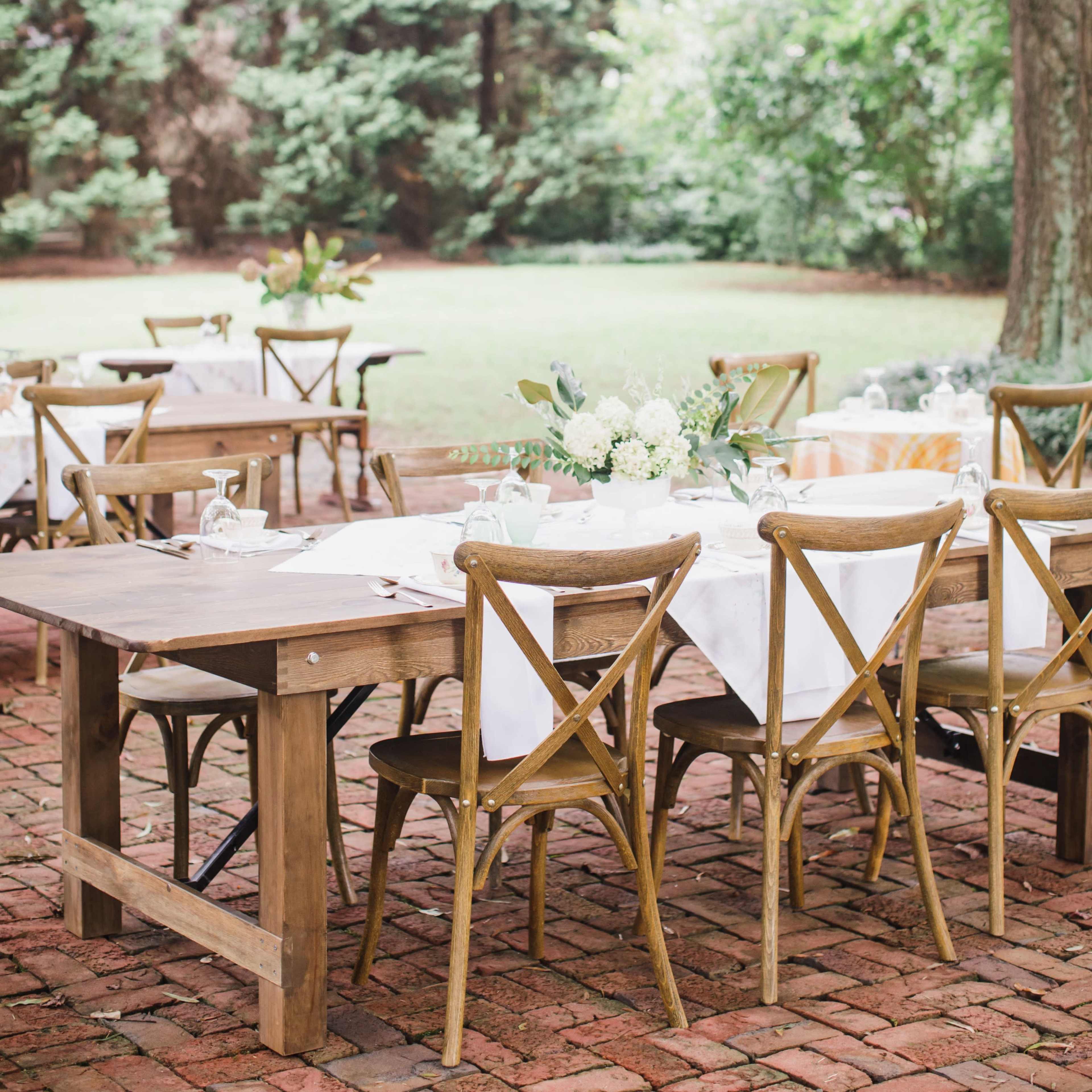 A rustic outdoor dining area features wooden tables and chairs set on a brick patio under a large tree, surrounded by greenery.