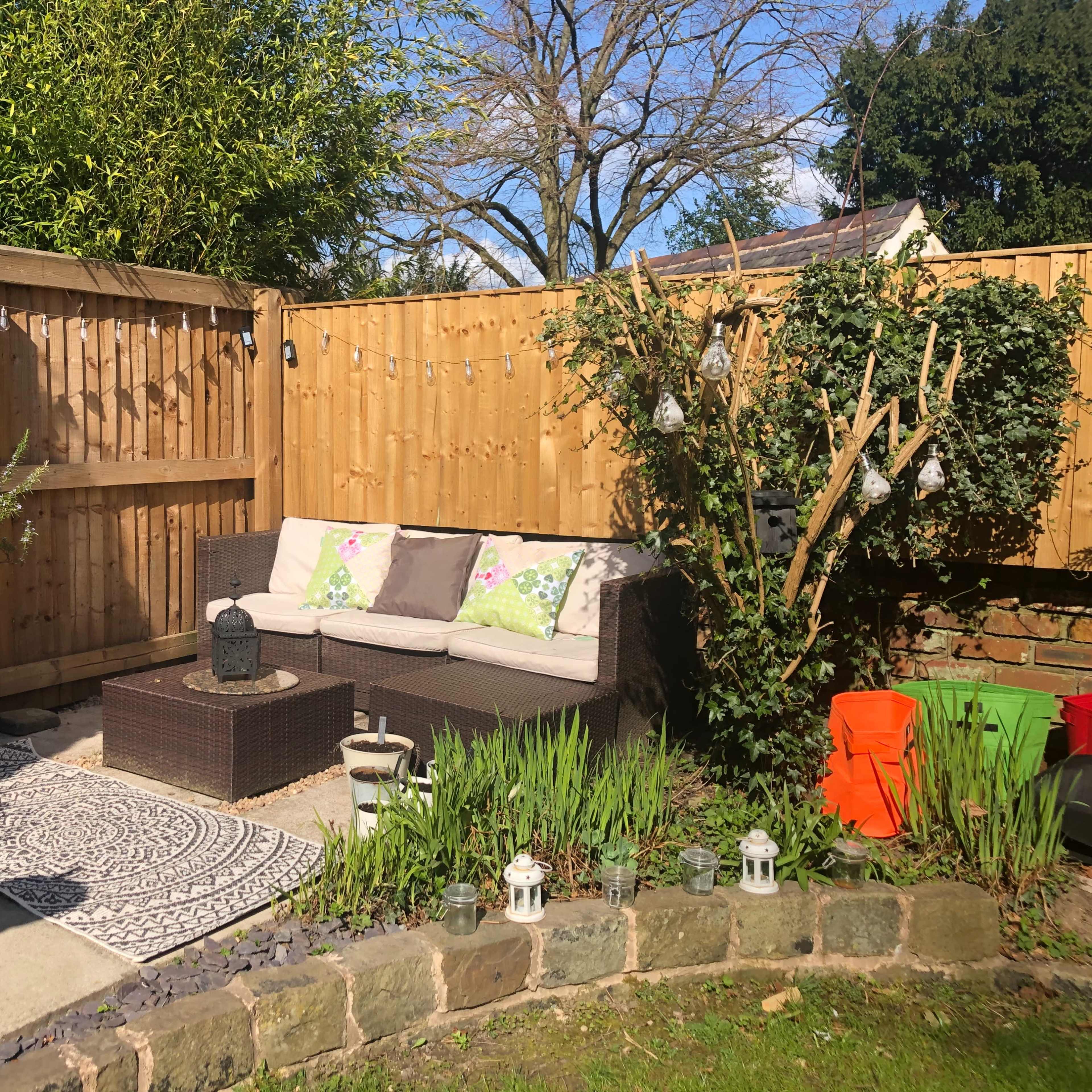 A small garden area featuring a seating arrangement with cushions, surrounded by plants and decorative elements like lanterns and garden containers.