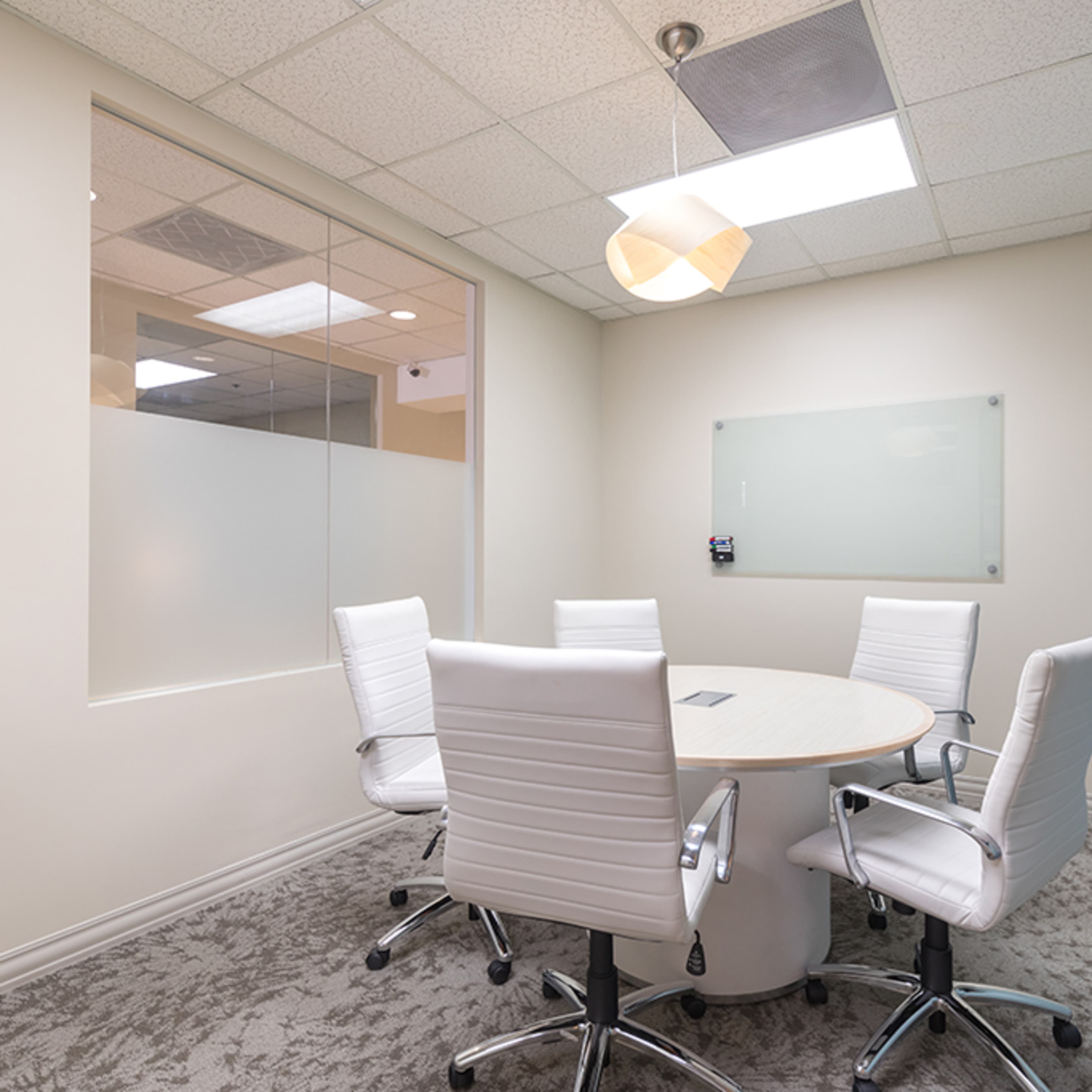 A small, modern conference room with a round table surrounded by five white ergonomic chairs.