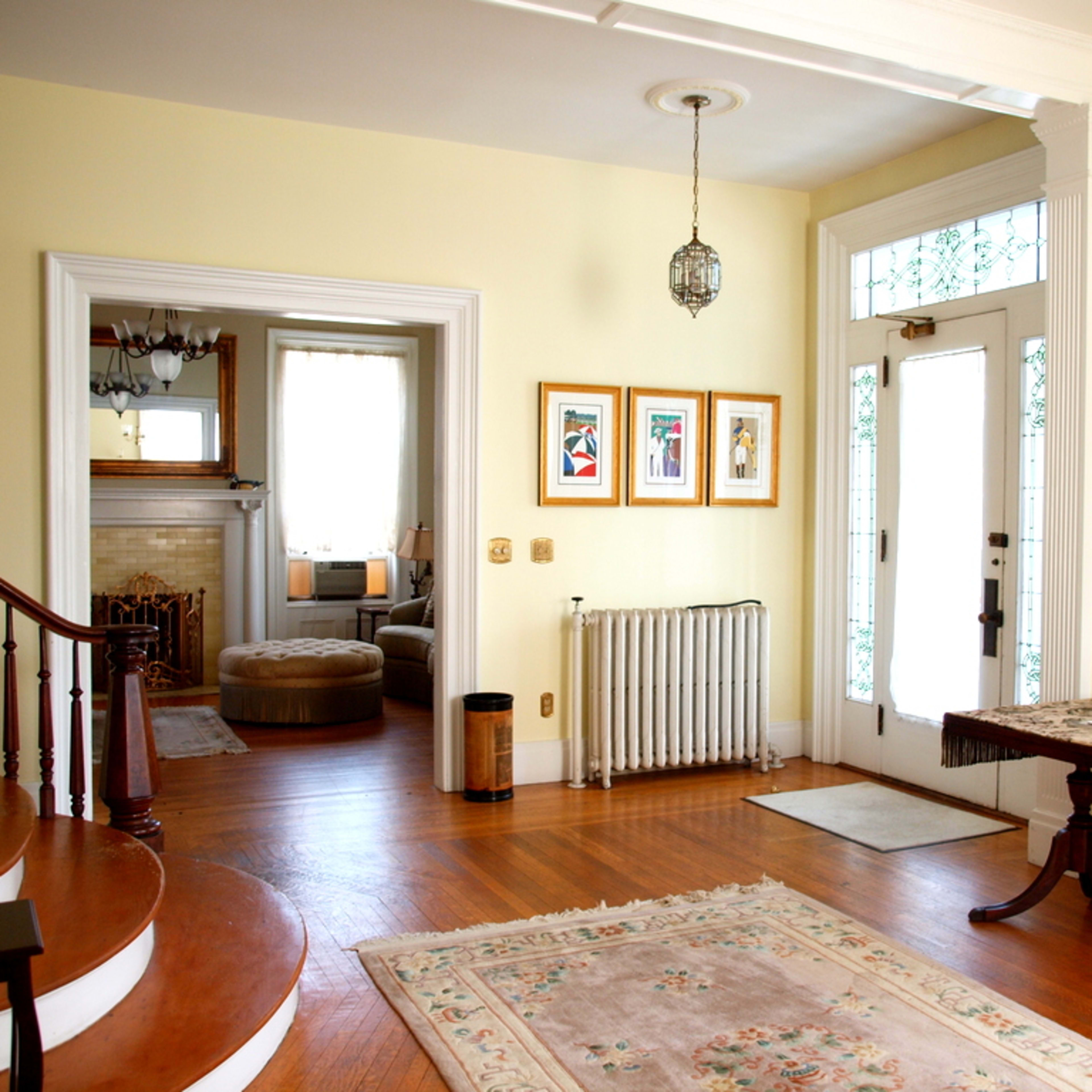A bright entryway with a staircase, framed art on the wall, a radiator, and a door leading to an adjacent room.