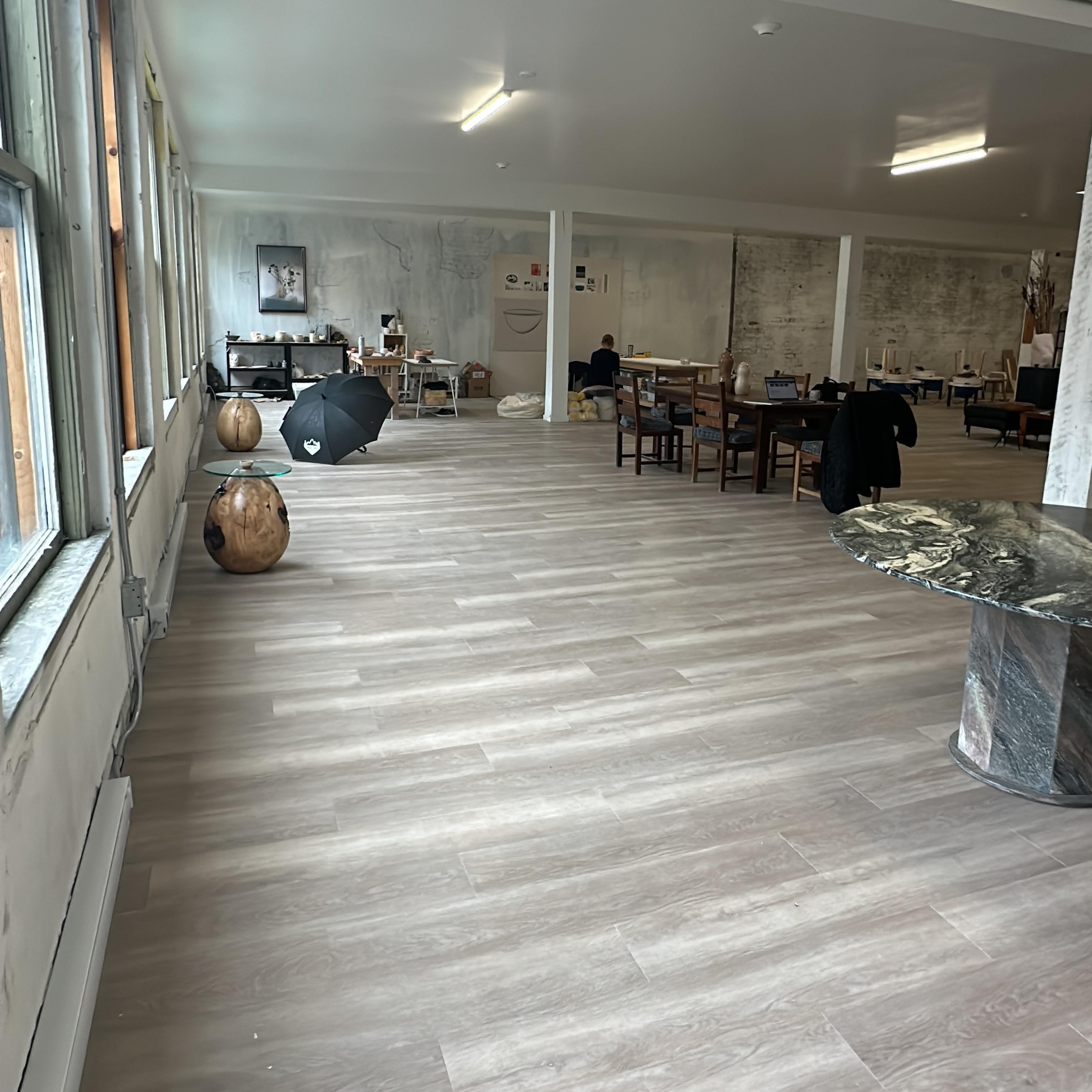 A spacious room with wooden flooring, large windows, a dining area with tables and chairs, and a few decorative objects.
