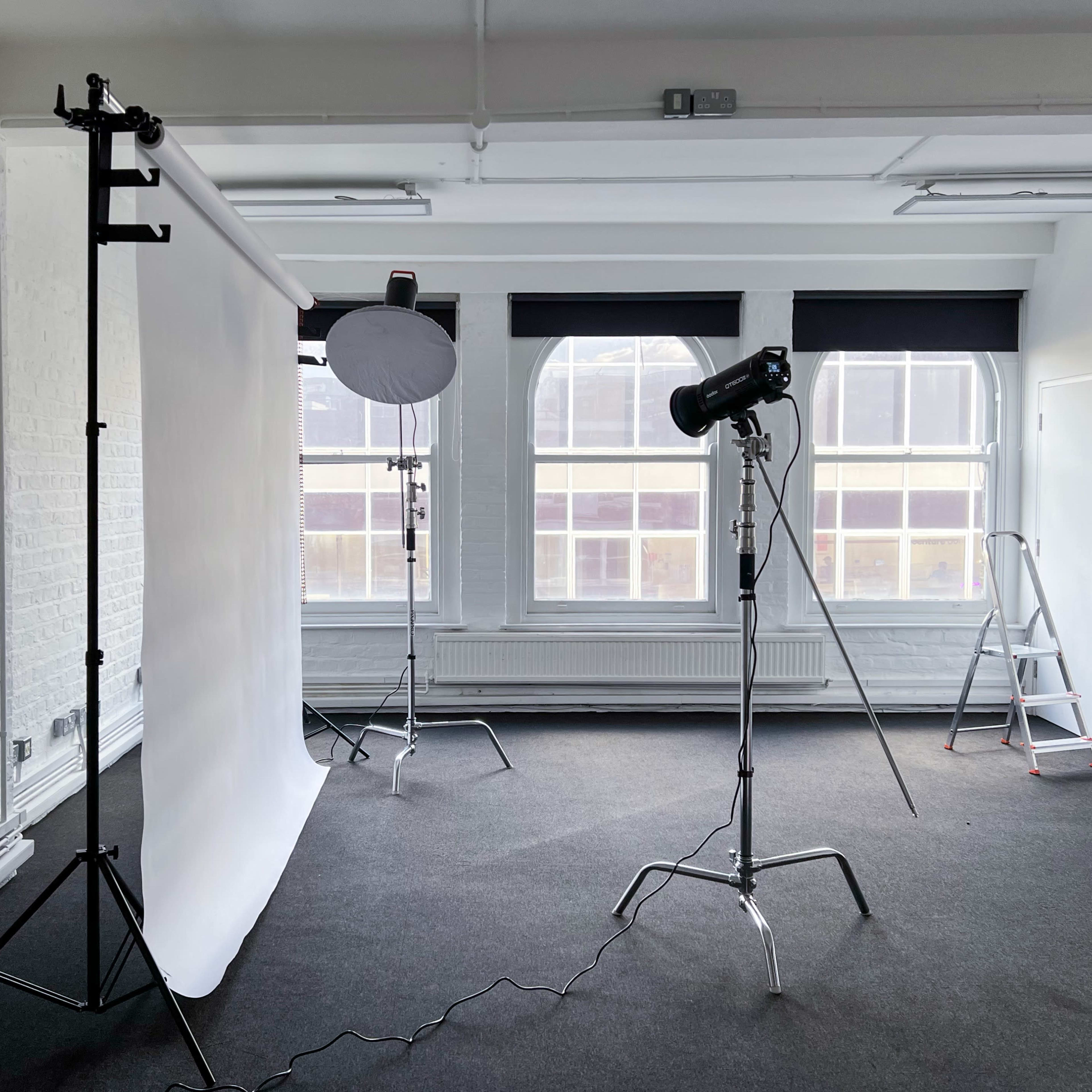 A photography studio features a white backdrop, lighting equipment, and a ladder in a spacious room with large windows.