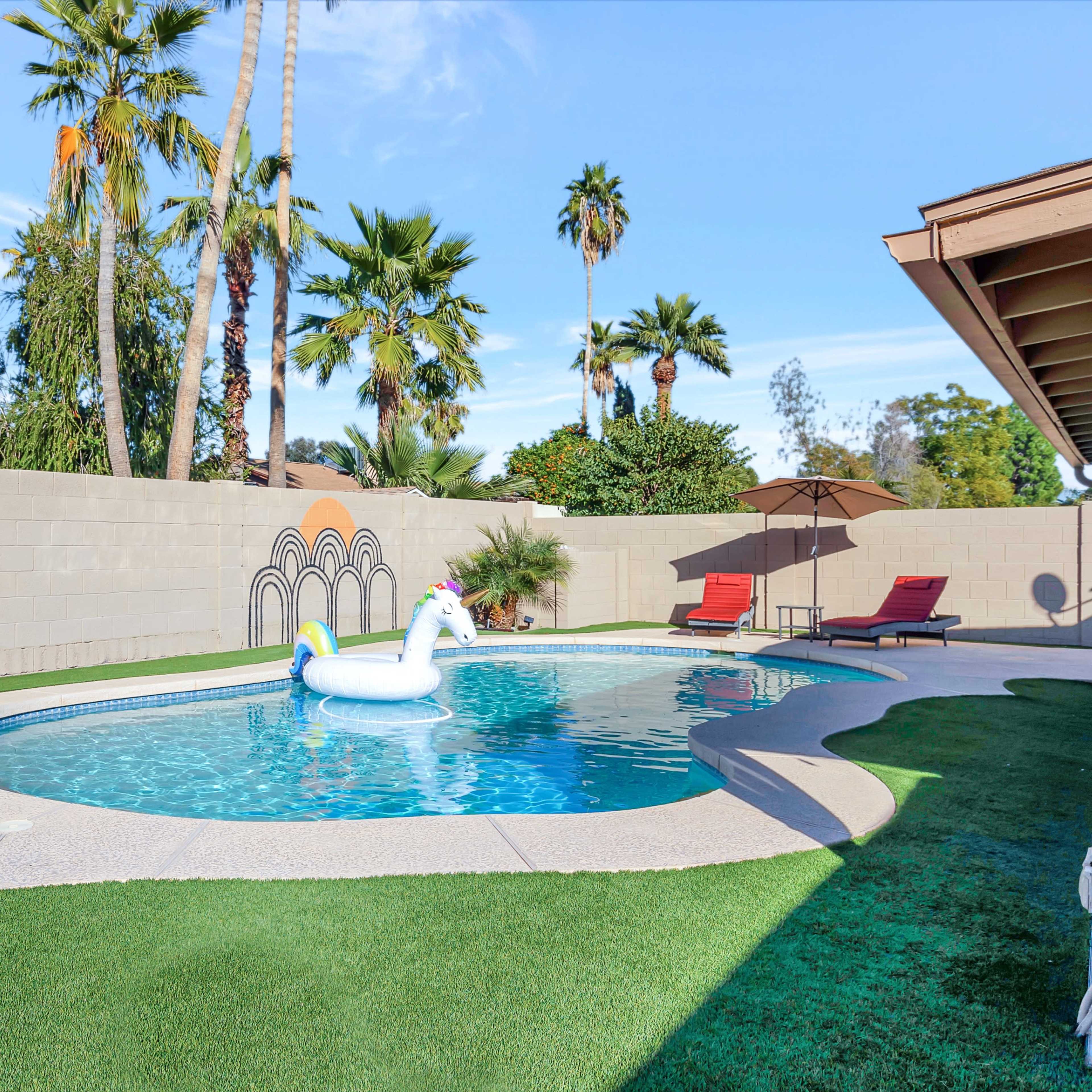 A backyard swimming pool with a unicorn float, surrounded by palm trees and two lounge chairs under a shade umbrella.