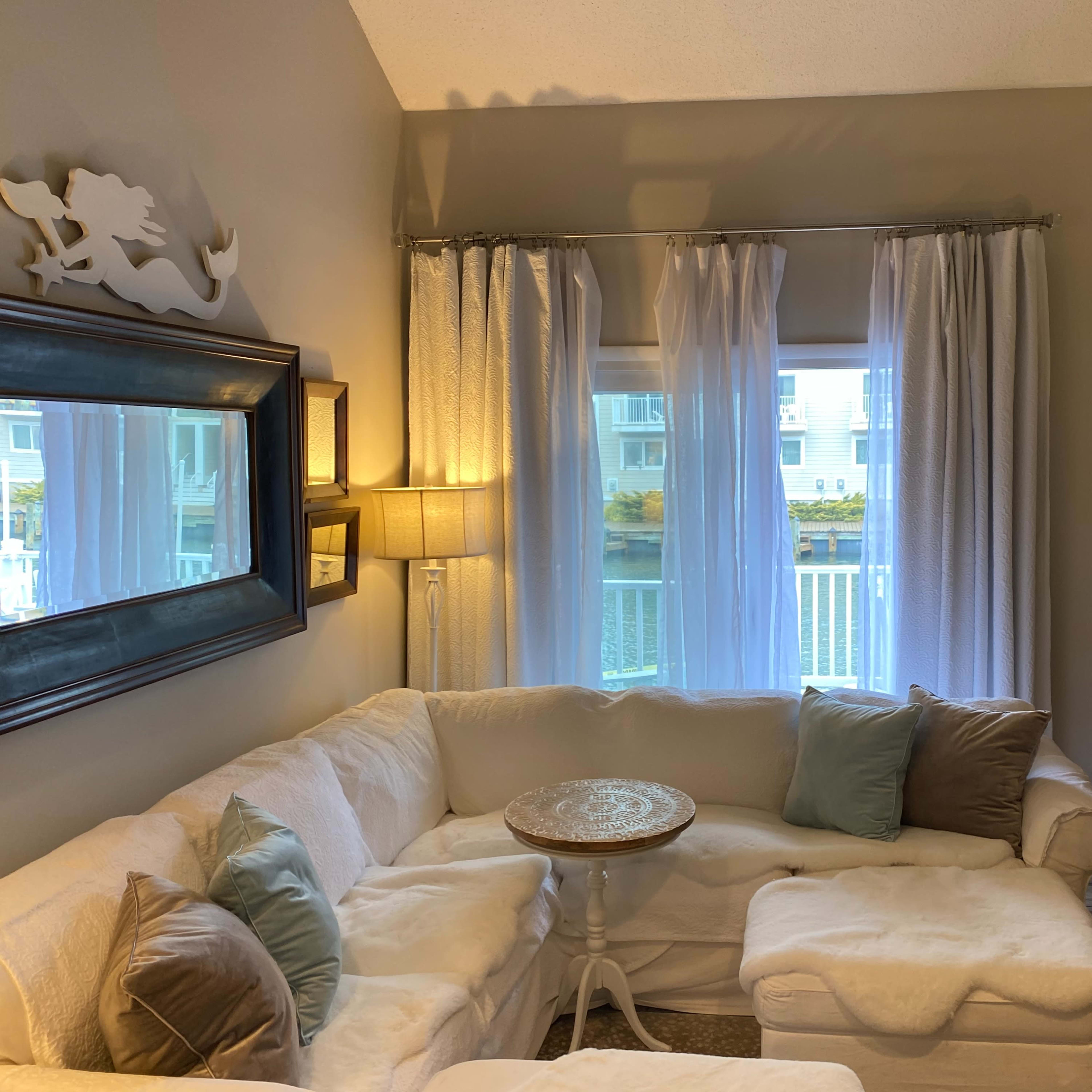 A cozy living room with a white sectional sofa, a small round table, and a fireplace illuminated by candles, all framed by sheer curtains and soft lighting.