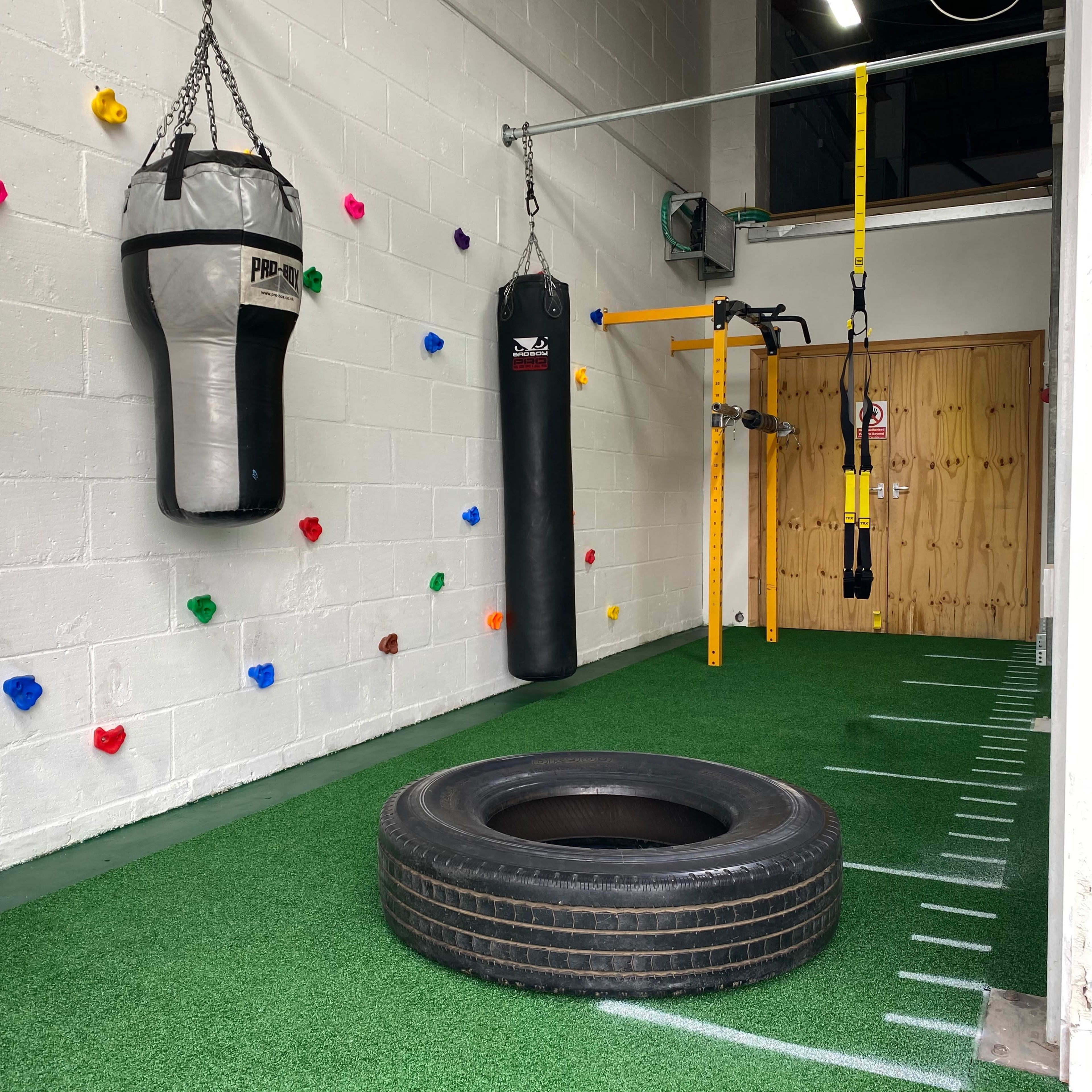 A small indoor workout space featuring a hanging punching bag, a battle rope setup, a tire on the green turf floor, and colorful climbing holds on the wall.