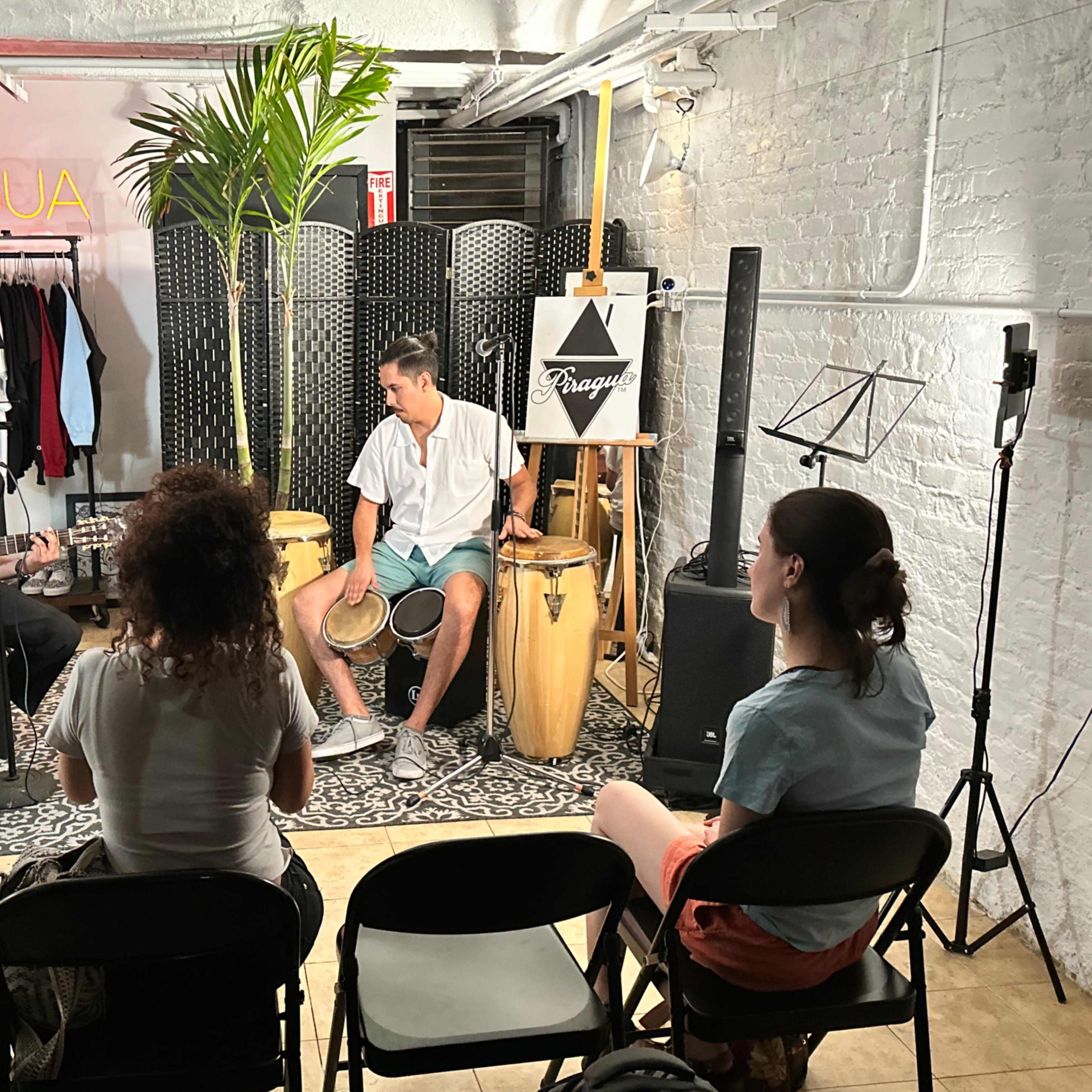 A musician plays guitar while another man drums, performing for an audience seated on black chairs in a small, decorated room.