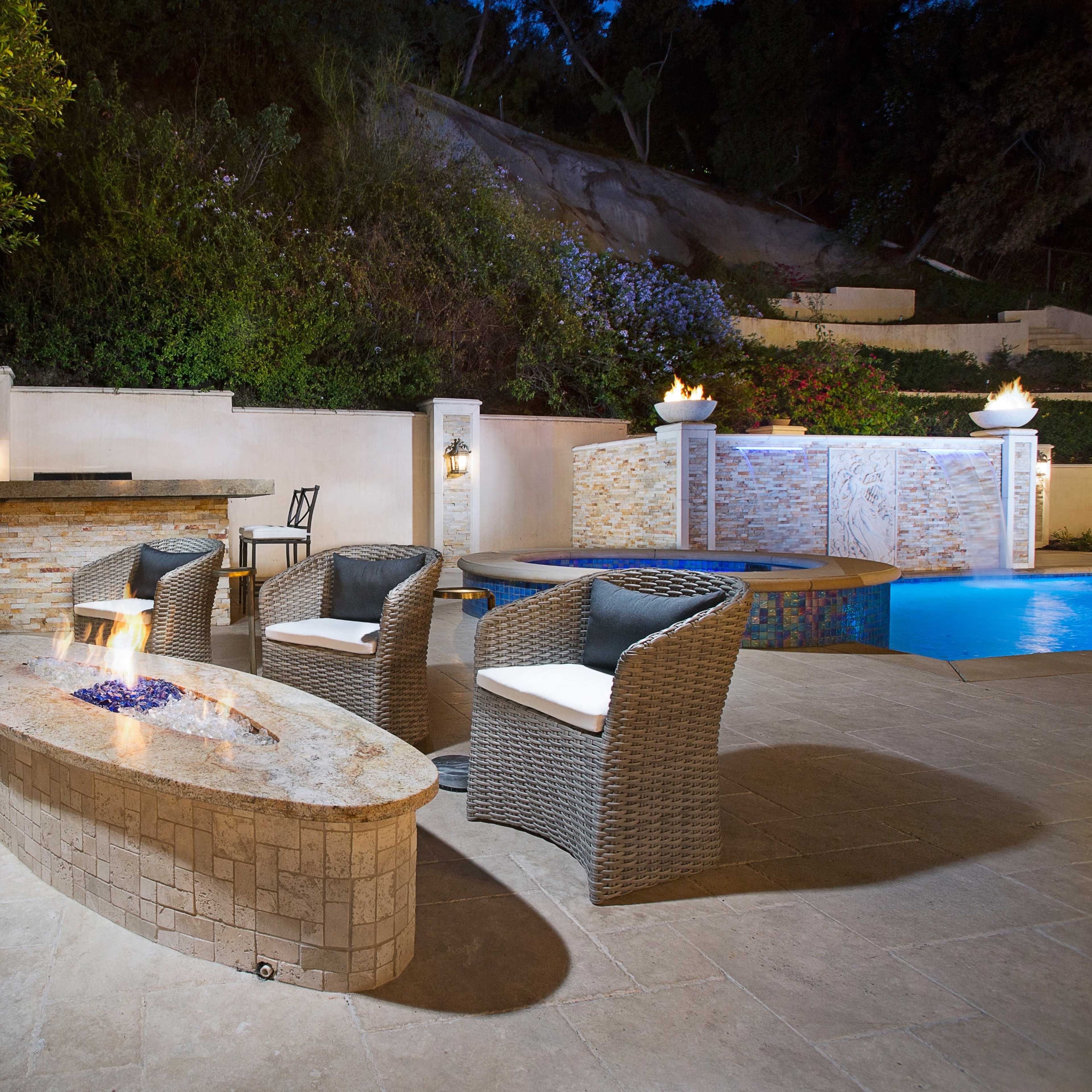 A modern outdoor patio area featuring a circular stone fire pit surrounded by four wicker chairs, with a swimming pool and landscaped greenery in the background.