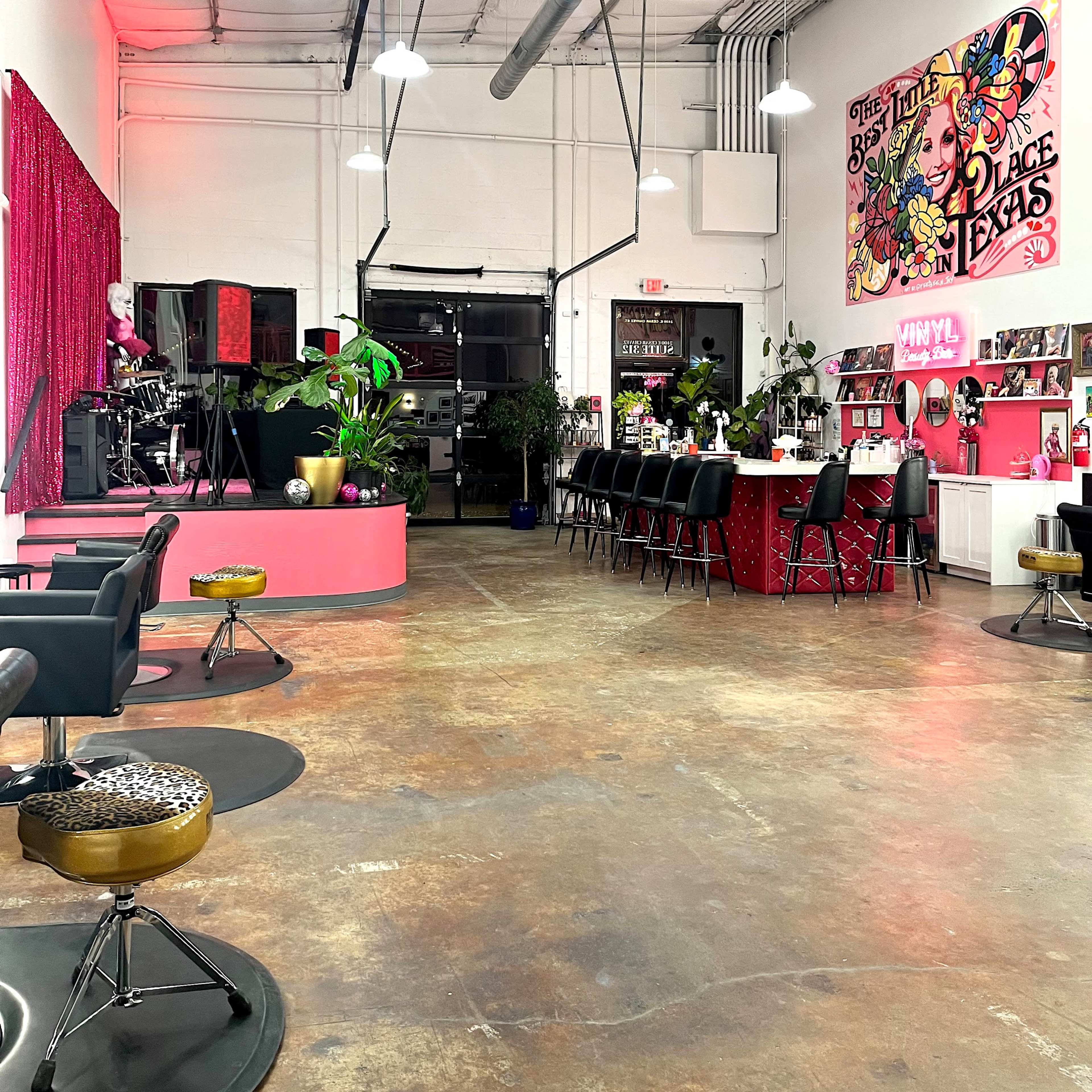 A brightly lit hair salon featuring a mix of pink and black furniture, with a reception area and styling stations arranged against the white walls.