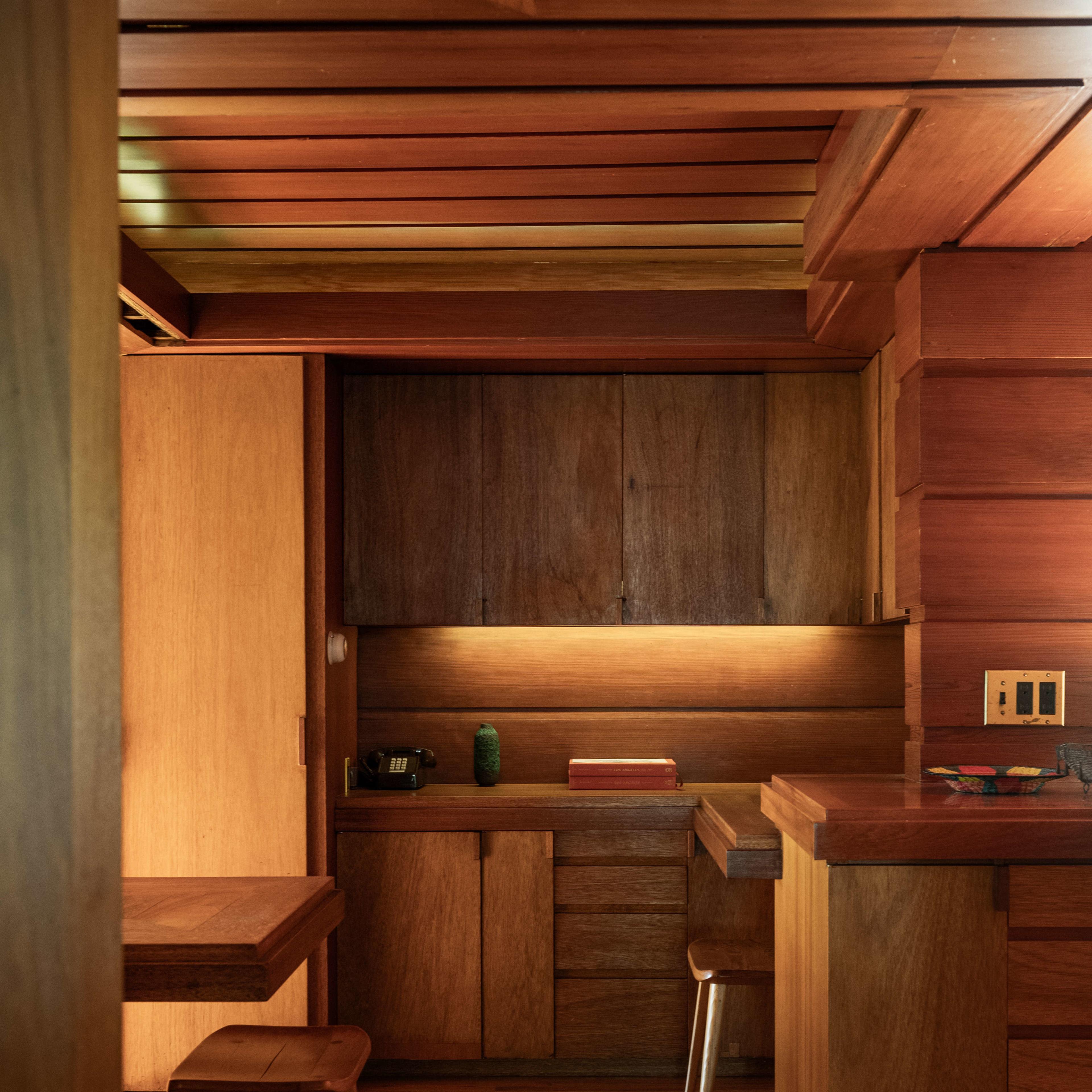 A wooden interior of a modern kitchen with a table, stools, and low cabinetry under warm lighting.