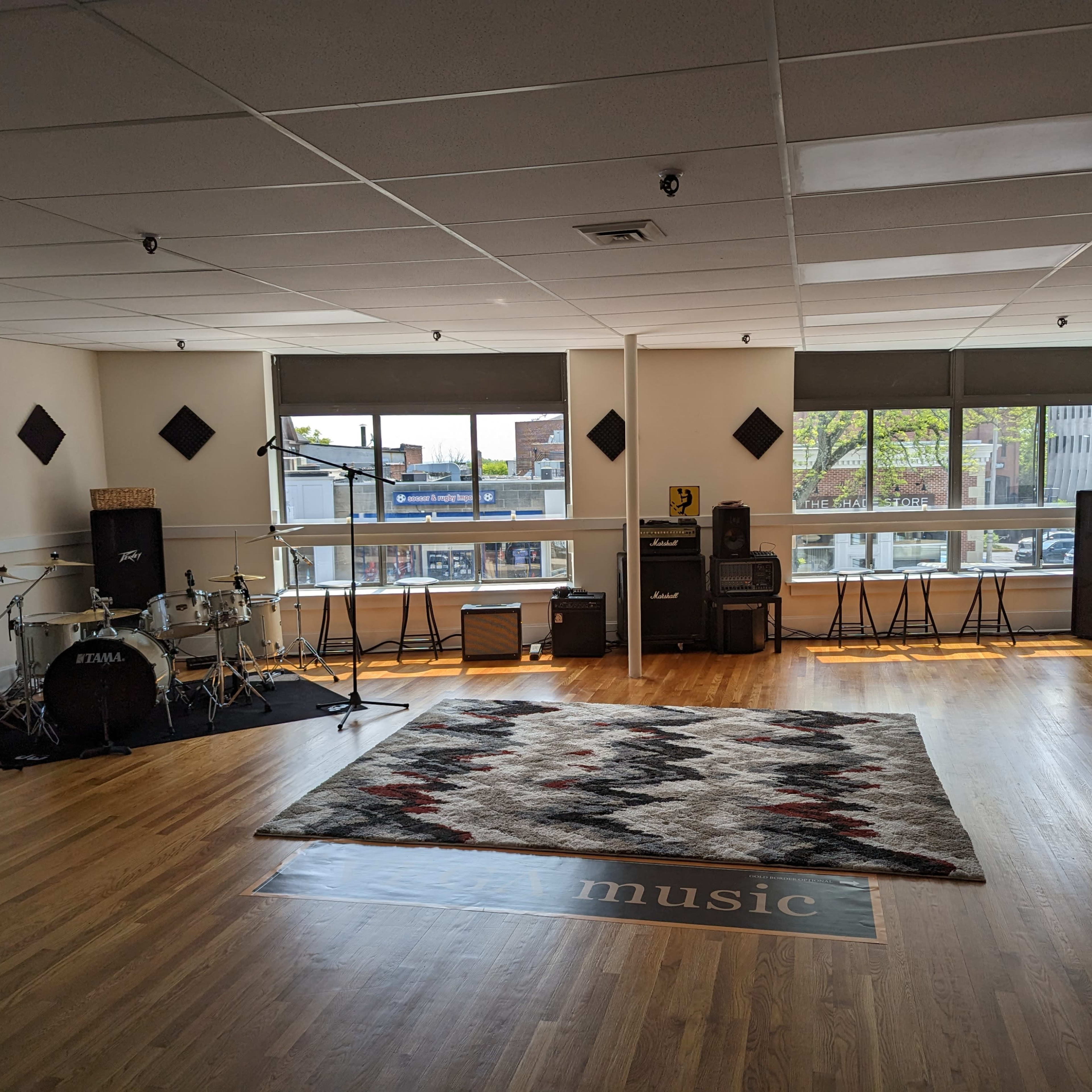A spacious music studio with a wooden floor, drum set, amplifiers, and a patterned rug centrally placed.