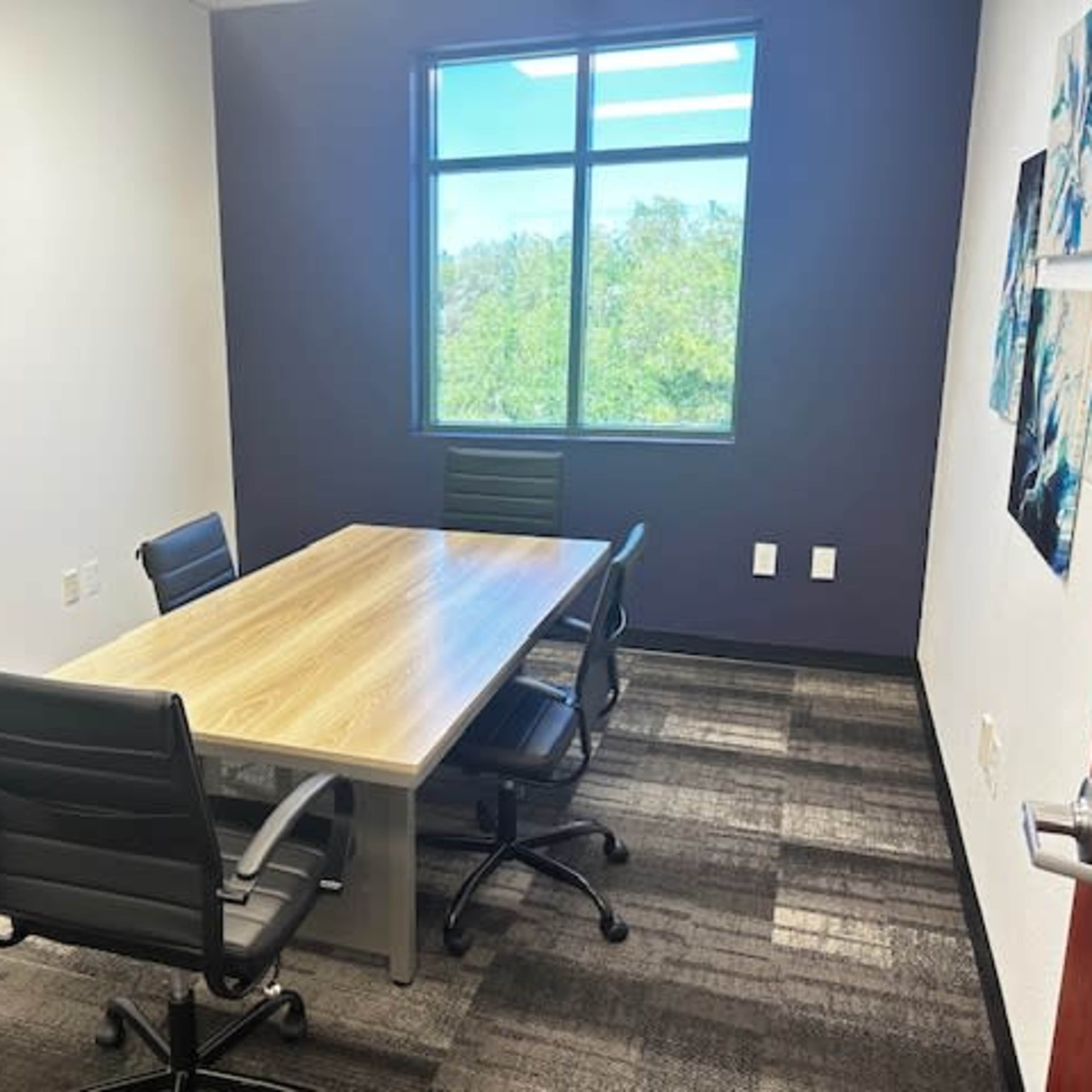 A conference room features a wooden table surrounded by four black chairs, with a wall-mounted TV and large window offering a view of greenery outside.