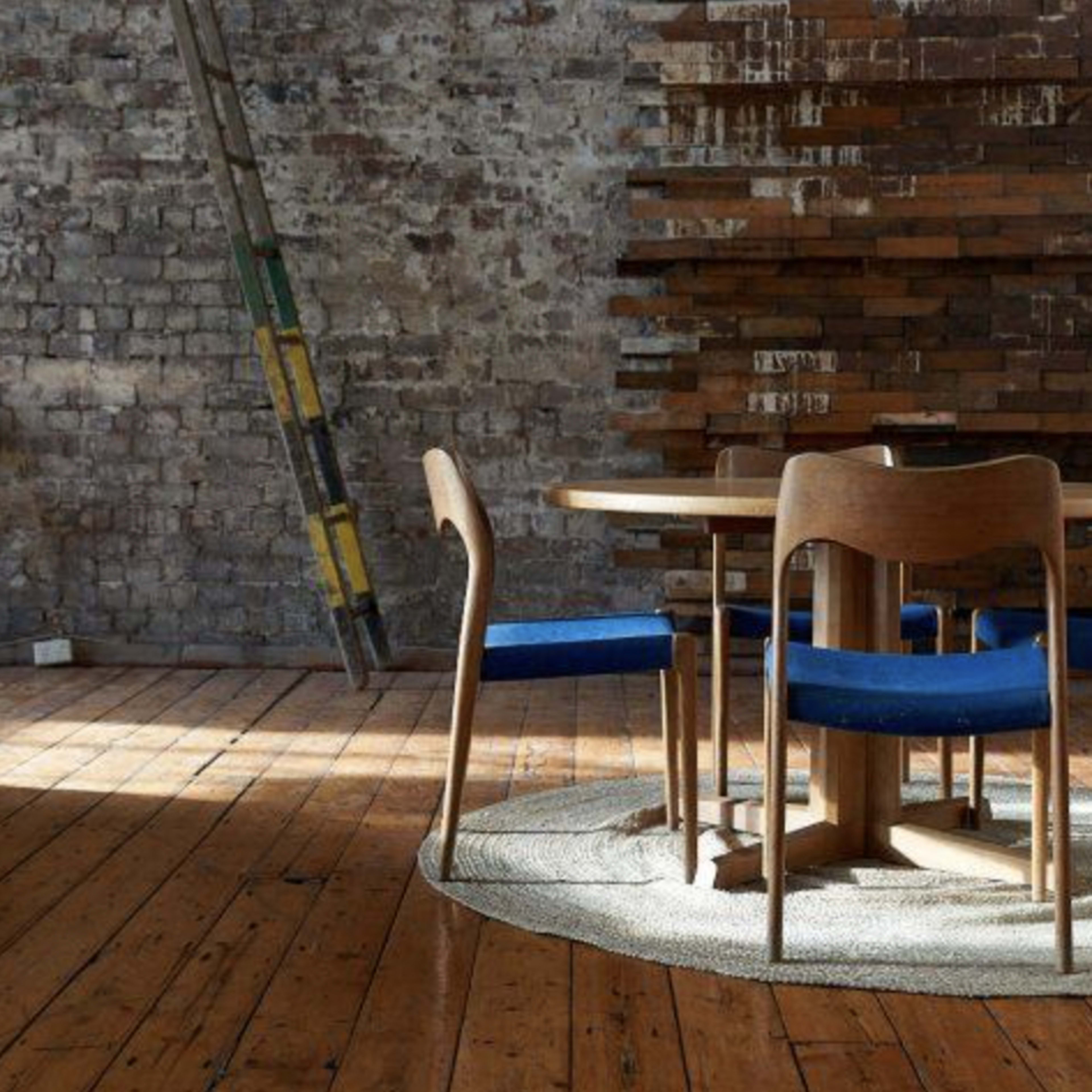 A round wooden table with blue chairs sits on a circular rug in a room with exposed brick walls and wooden flooring.