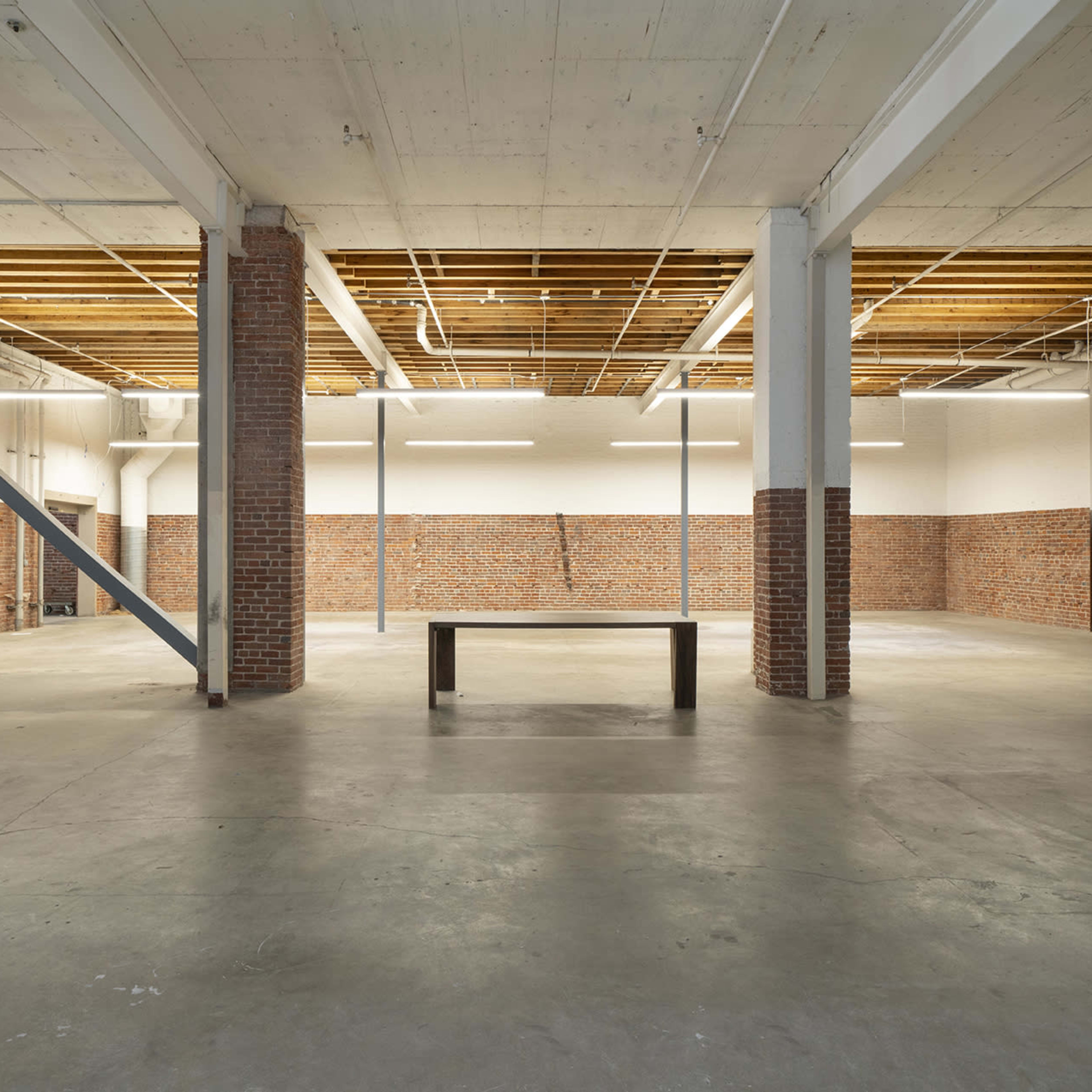 A spacious, empty industrial interior with concrete floors, exposed brick walls, and a central table supported by metal beams.