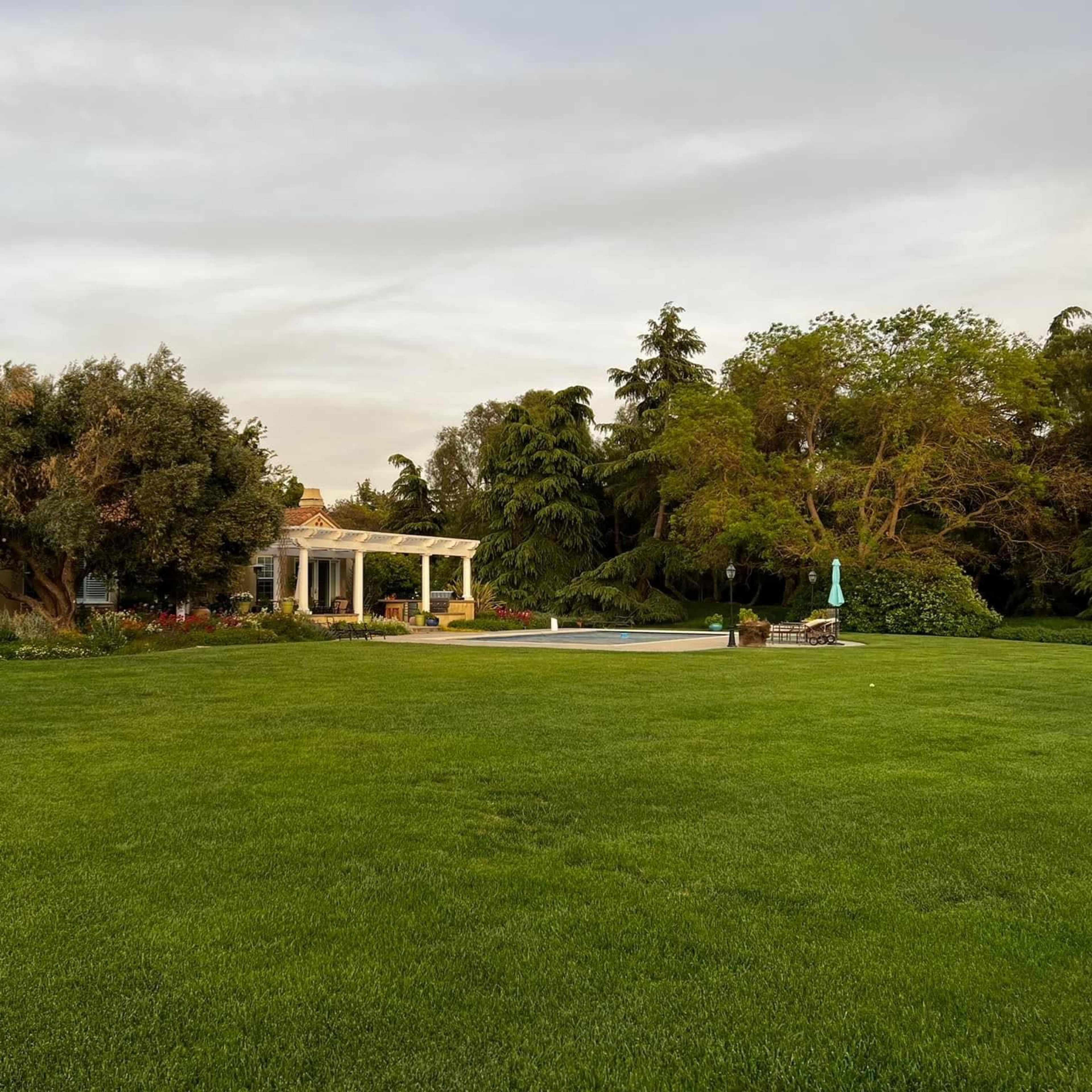 A single-story house with a covered patio and a swimming pool is set in a spacious green lawn surrounded by trees and colorful landscaping.